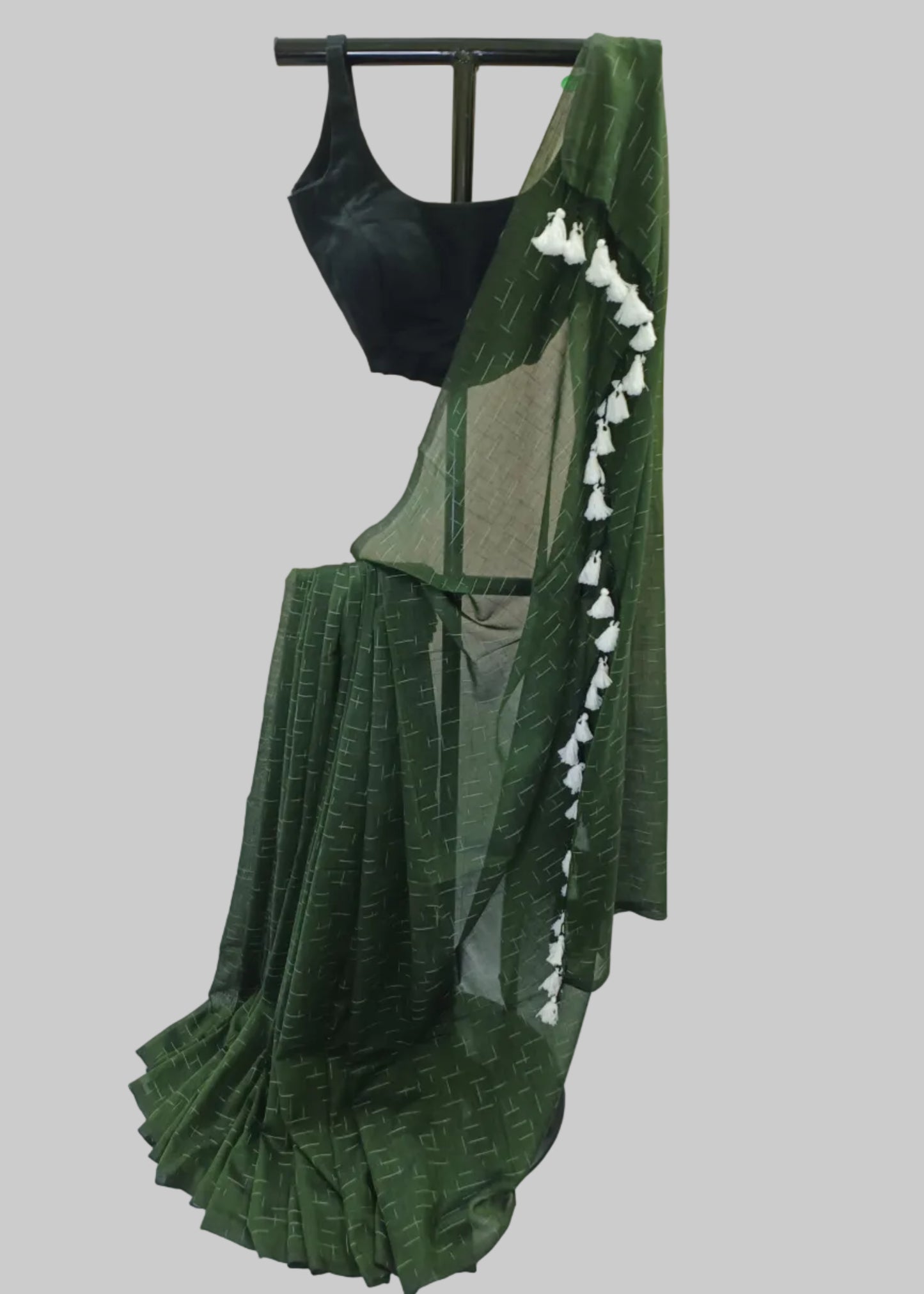 A dark green saree with a subtle geometric pattern, draped on a black dress form. The saree features a black sleeveless blouse and a green border adorned with white tassels, set against a draped white fabric backdrop.
