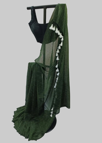 A dark green saree with a subtle geometric pattern, draped on a black dress form. The saree features a black sleeveless blouse and a sheer green pallu adorned with white tassels, set against a draped white fabric backdrop.