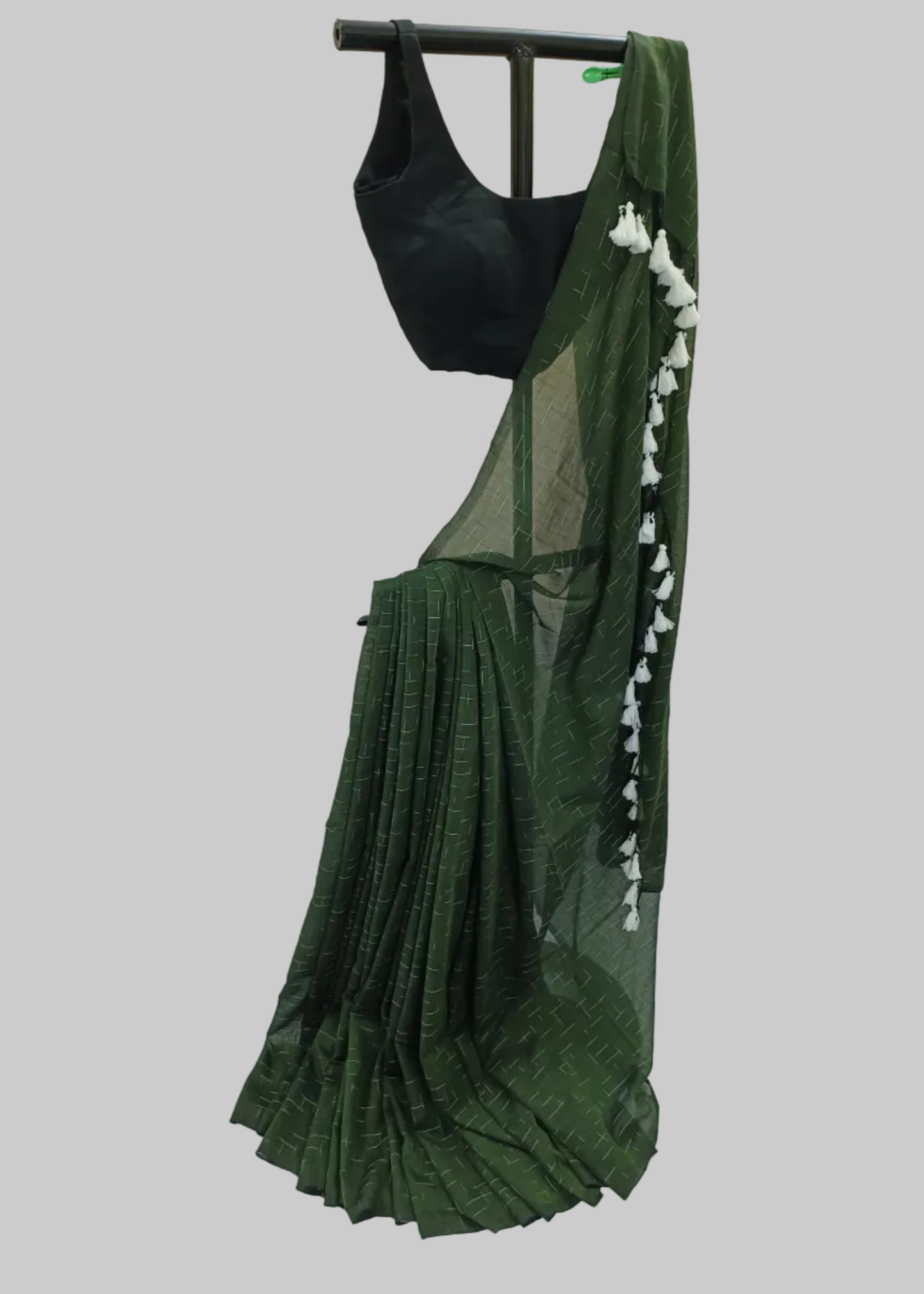 A dark green saree with a subtle woven pattern, draped over a black sleeveless blouse, hanging on a black clothing rack against a draped white fabric backdrop.