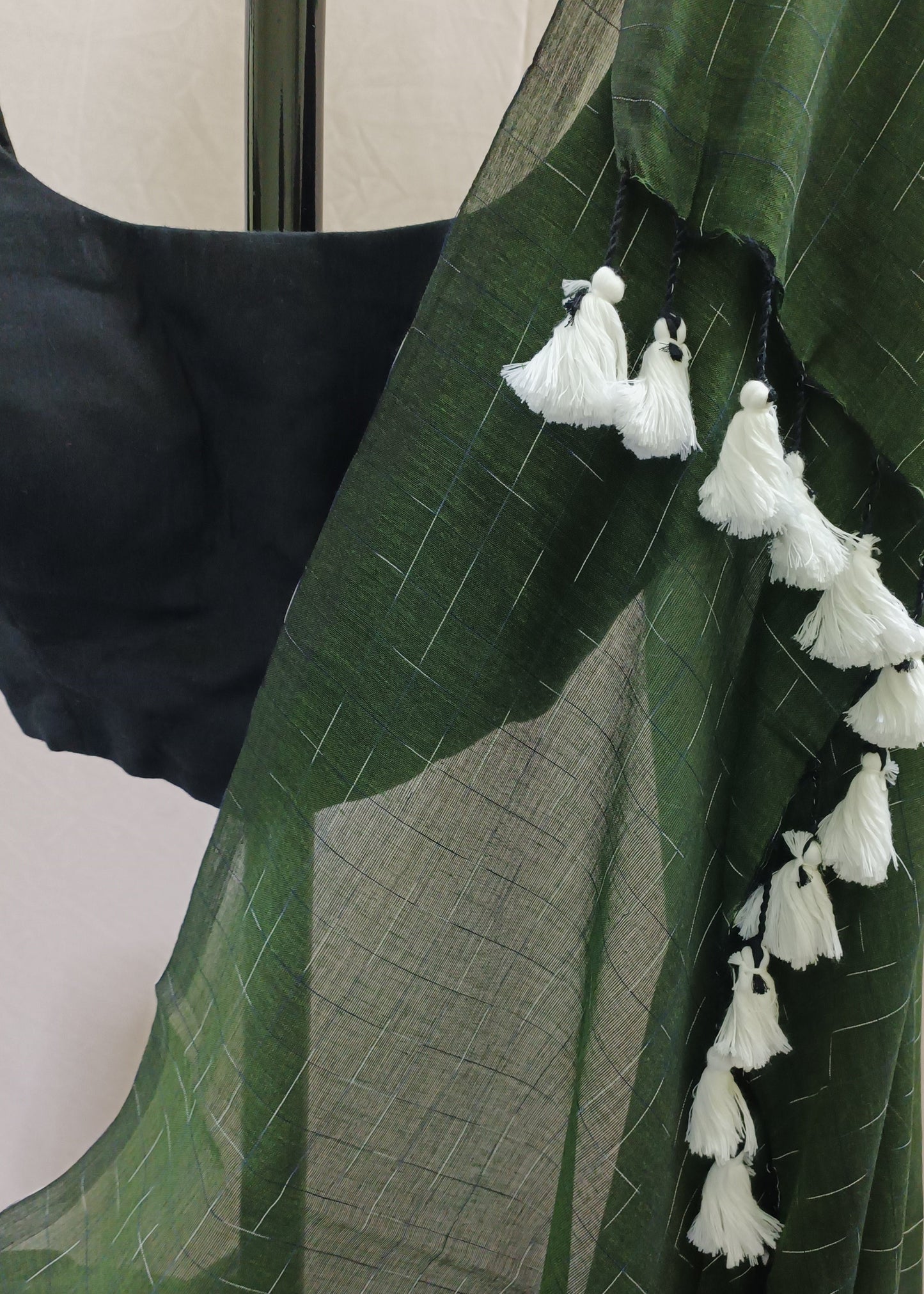 A dark green sheer saree drape with white tassels along the border, paired with a black blouse, shown against a neutral beige background.