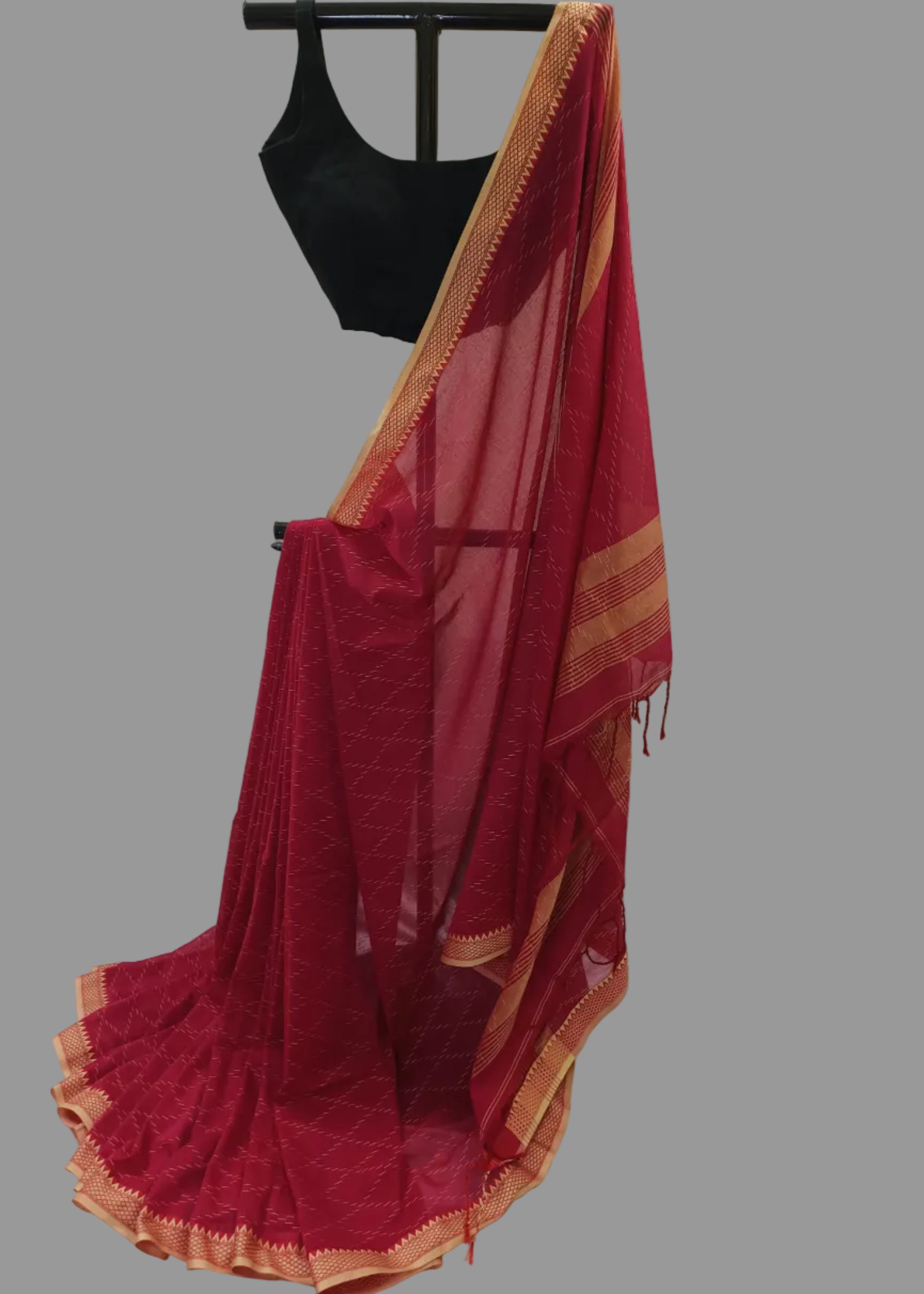 A deep red saree with gold zari borders and a matching black blouse displayed on a hanger against a draped white fabric backdrop.