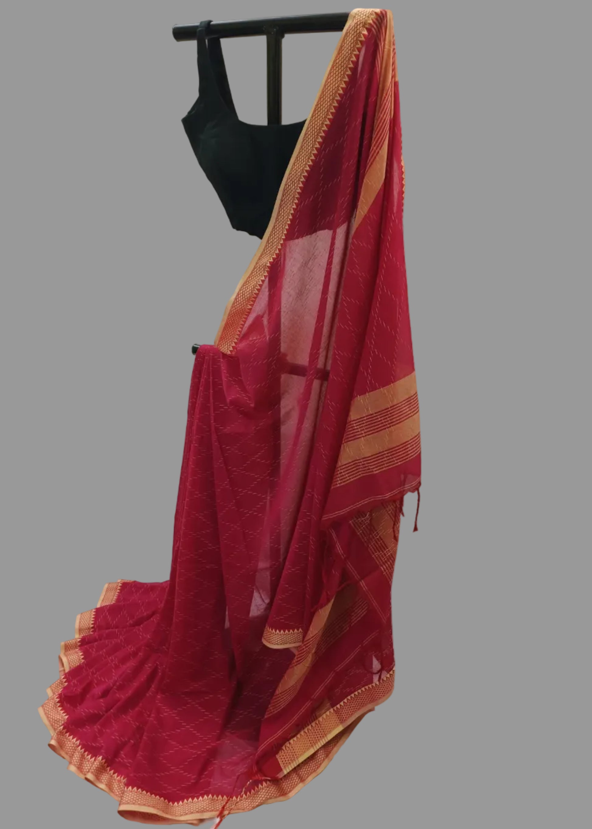 A deep red saree with a gold border and a matching red blouse displayed on a black hanger against a draped white fabric backdrop.