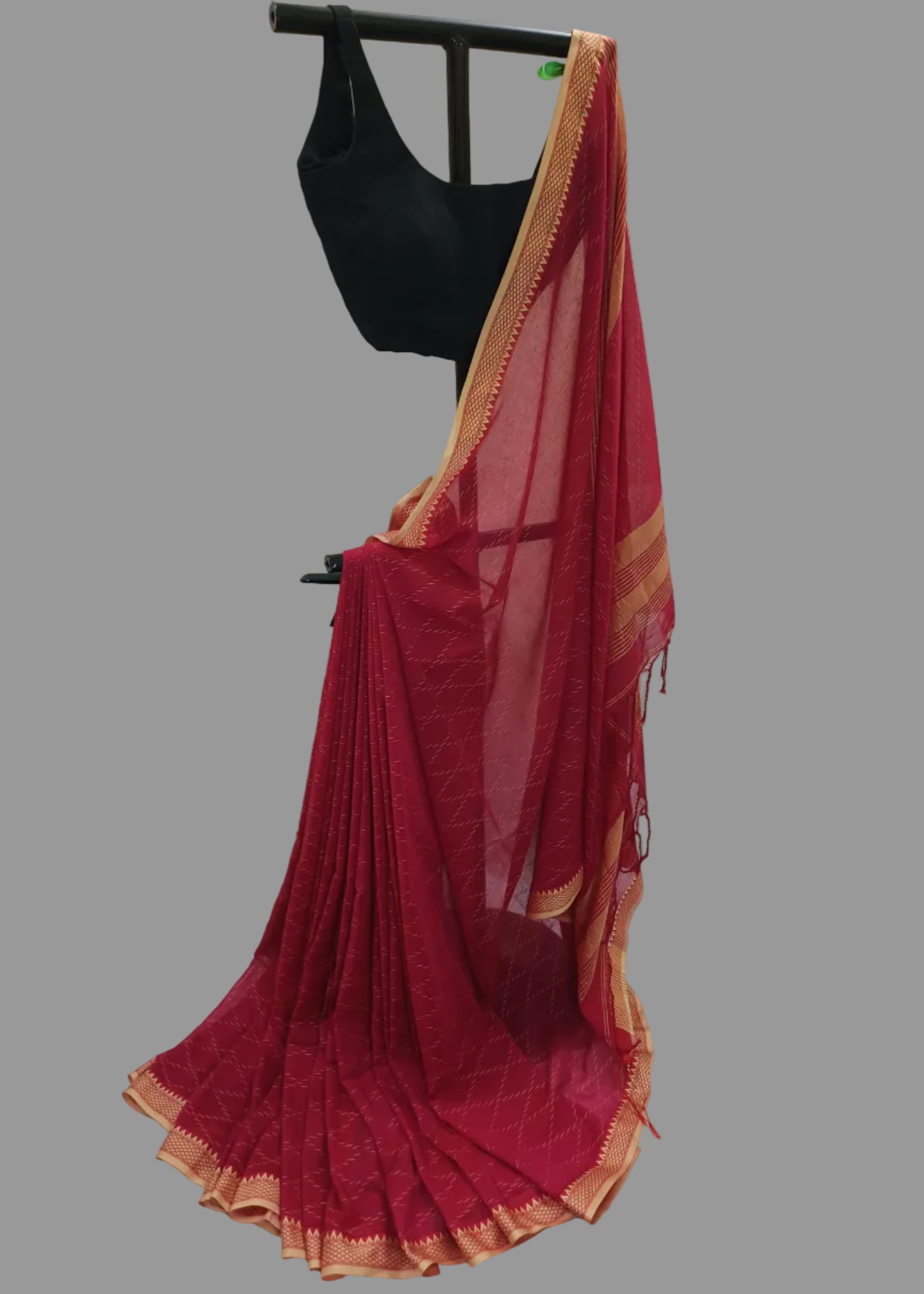 A deep red saree with gold zari borders draped on a black hanger stand, paired with a black sleeveless blouse, set against a white draped fabric backdrop.