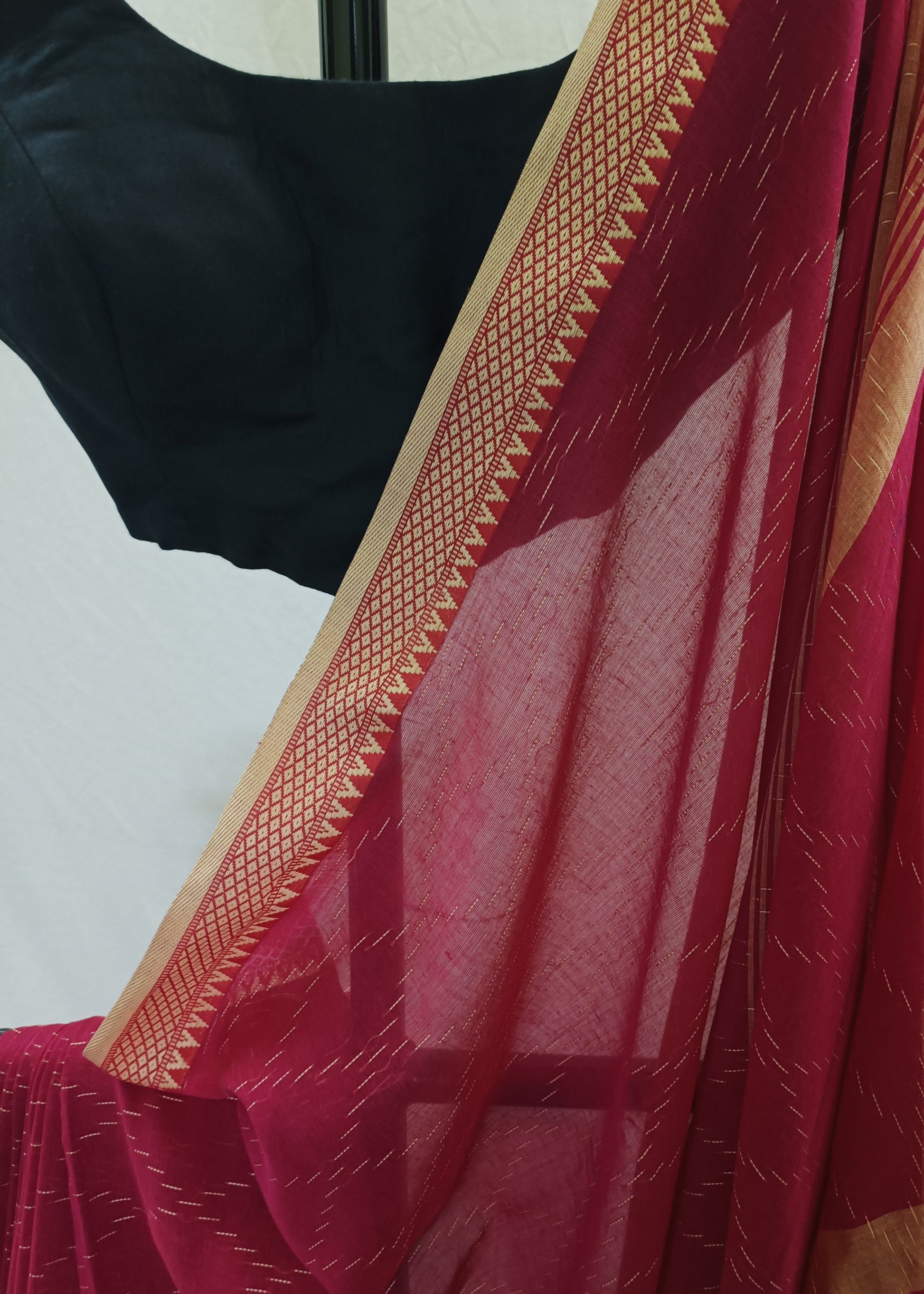 A close-up of a deep pinkish-red saree with a gold patterned border, draped over a black blouse against a light off-white fabric backdrop.
