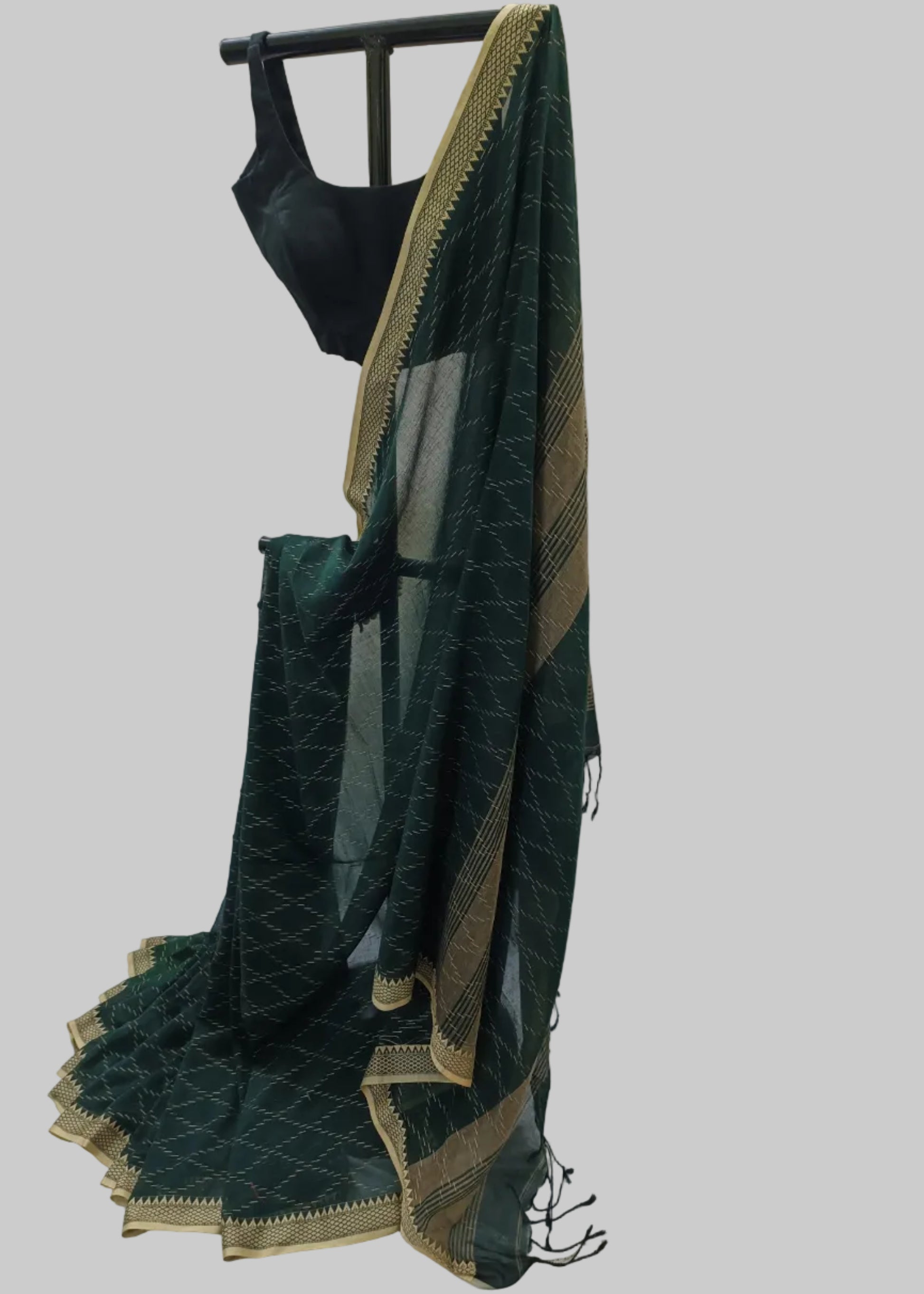 A dark green saree with gold zari geometric patterns and borders, draped on a black clothing stand with a black blouse, set against a draped white fabric backdrop.