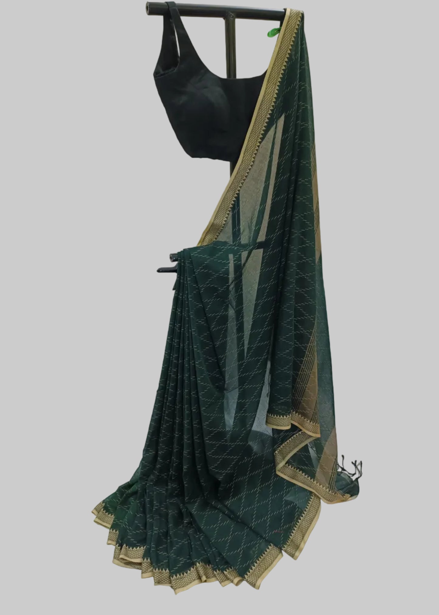 A dark green saree with a gold border and geometric pattern, paired with a black blouse, displayed on a clothing hanger against a draped white fabric backdrop.