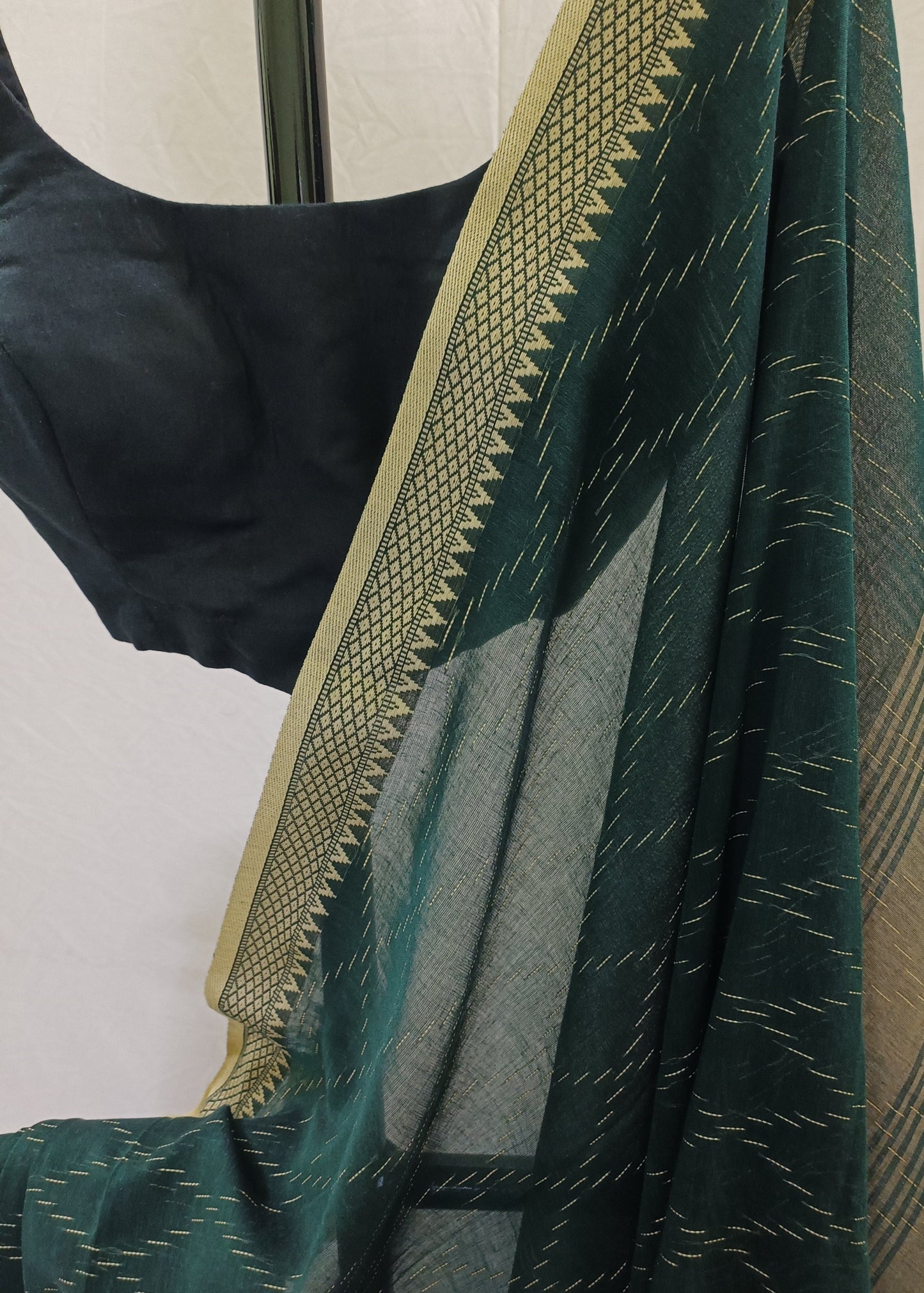 A dark green saree with gold zari ikat patterns and a wide gold border, draped over a black blouse against a light off-white background.