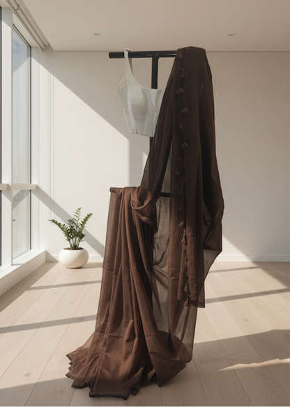Brown mul cotton saree hanging on a rack in a bright room with large windows.