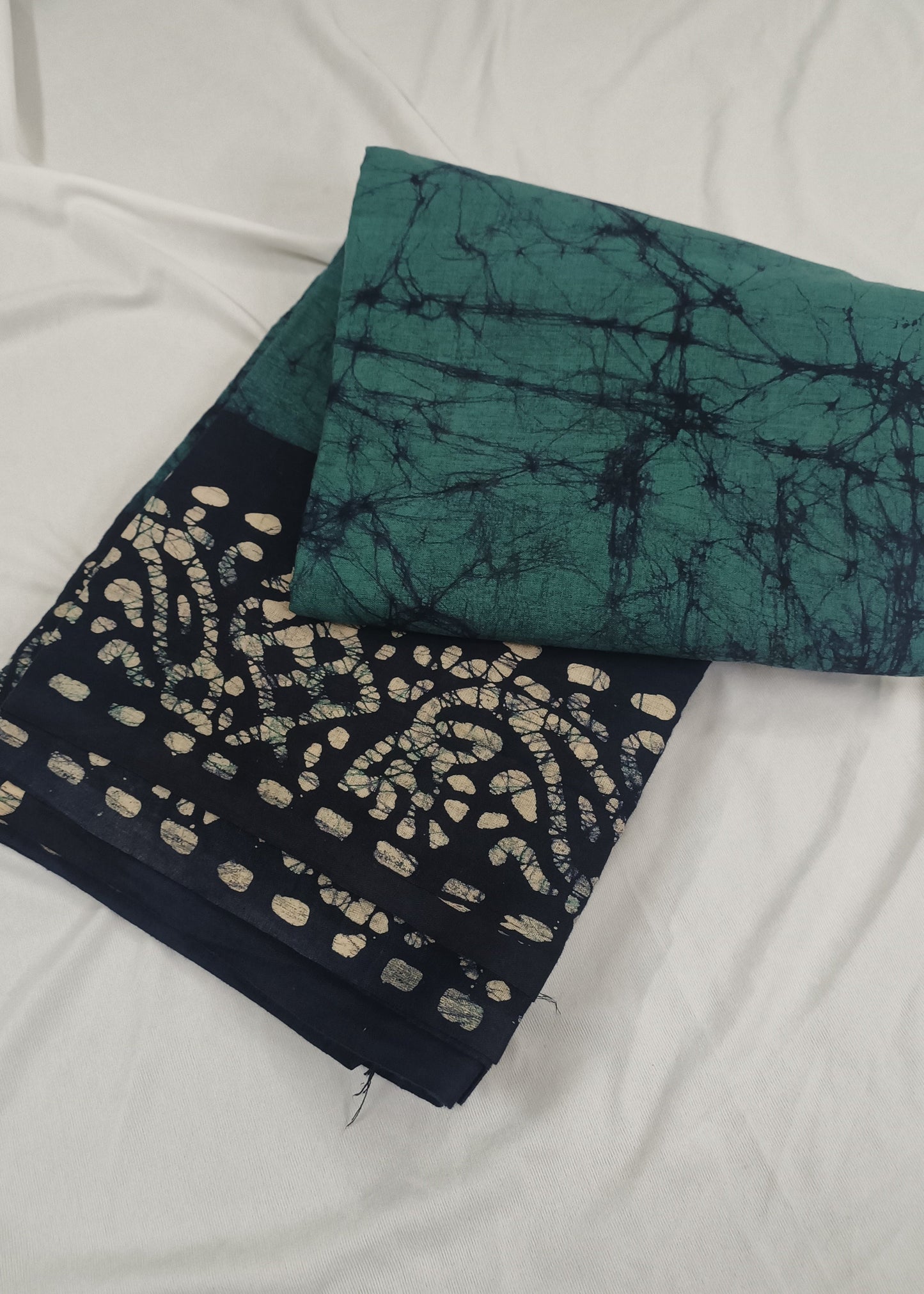 Two batik patterned cloths, one green and one black with white patterns, on a white fabric background.