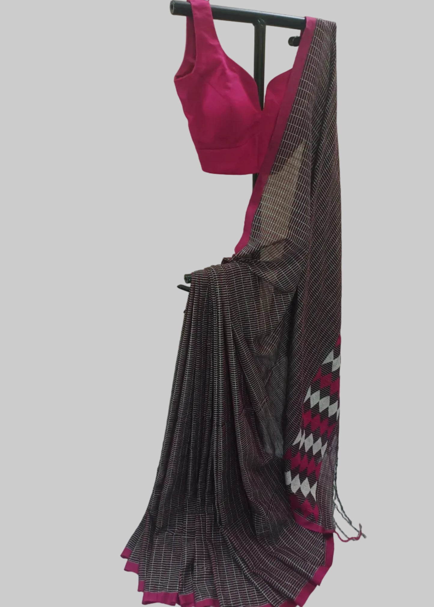 A dark gray and black patterned saree with a magenta pink blouse and border, displayed on a black clothing rack against a draped cream fabric backdrop.