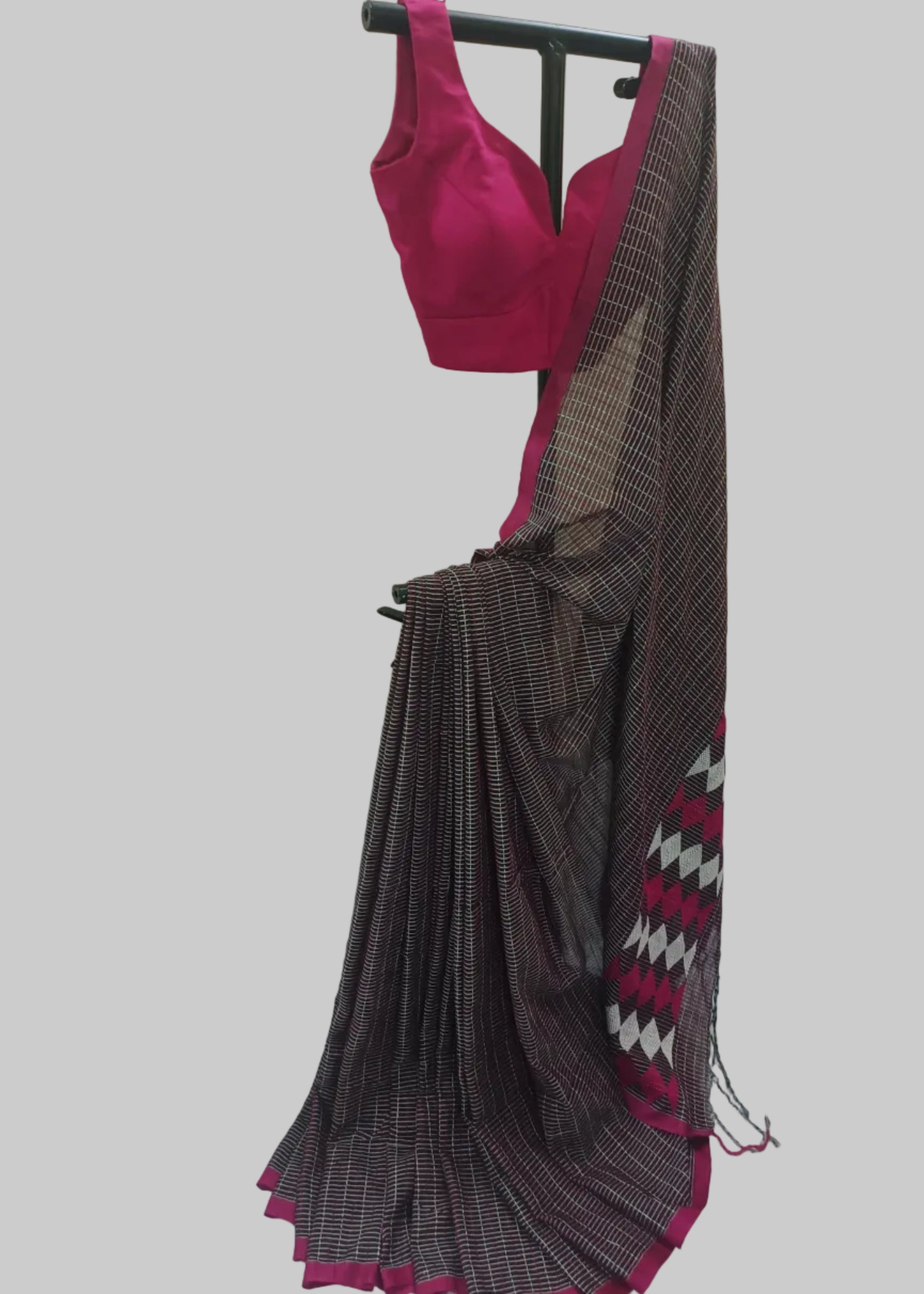 A dark gray and black patterned saree with a magenta pink blouse and border, displayed on a black clothing rack against a draped cream fabric backdrop.