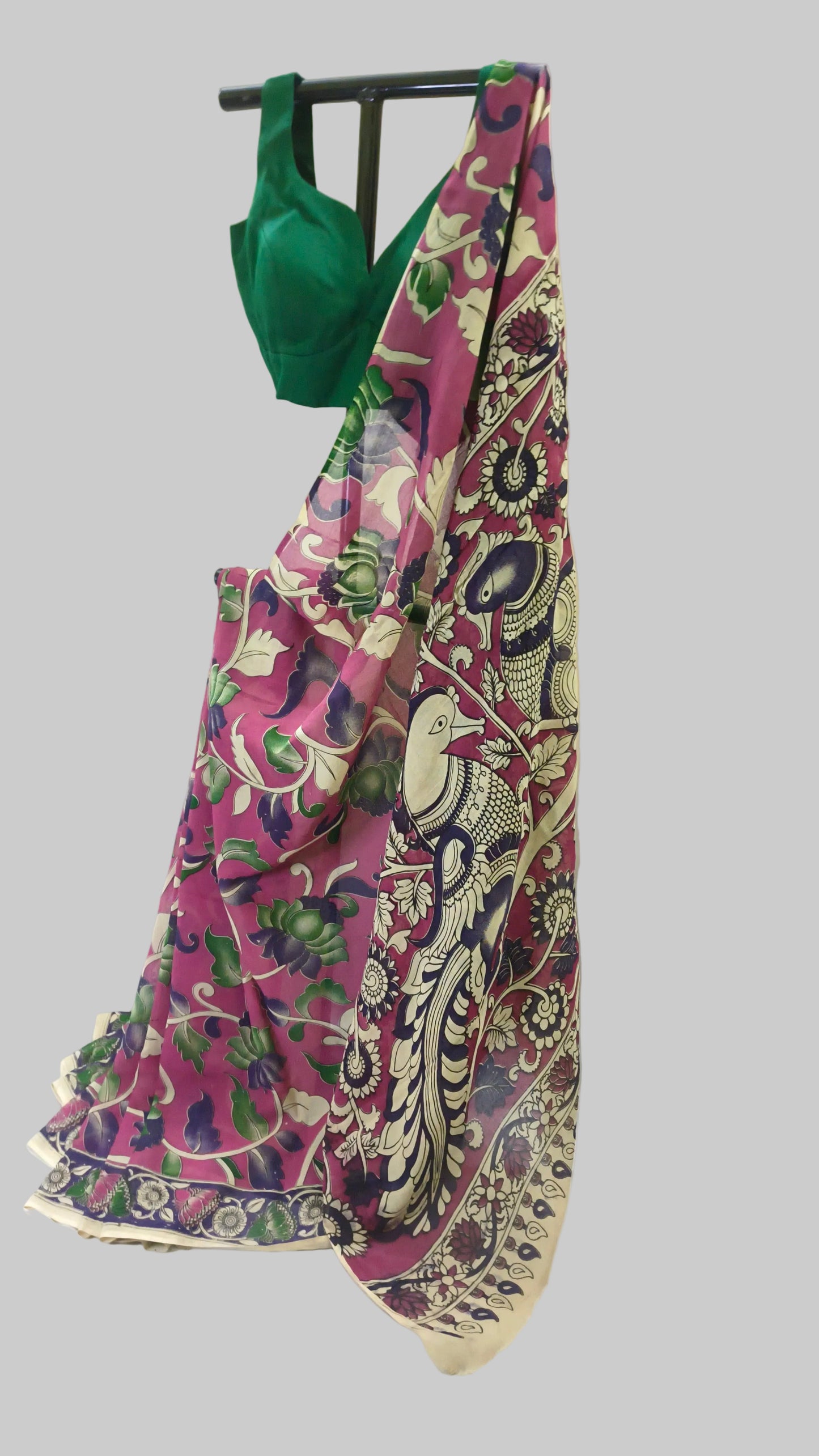 A pink and multicolor printed saree with a green blouse draped over a black hanger against a white fabric backdrop.