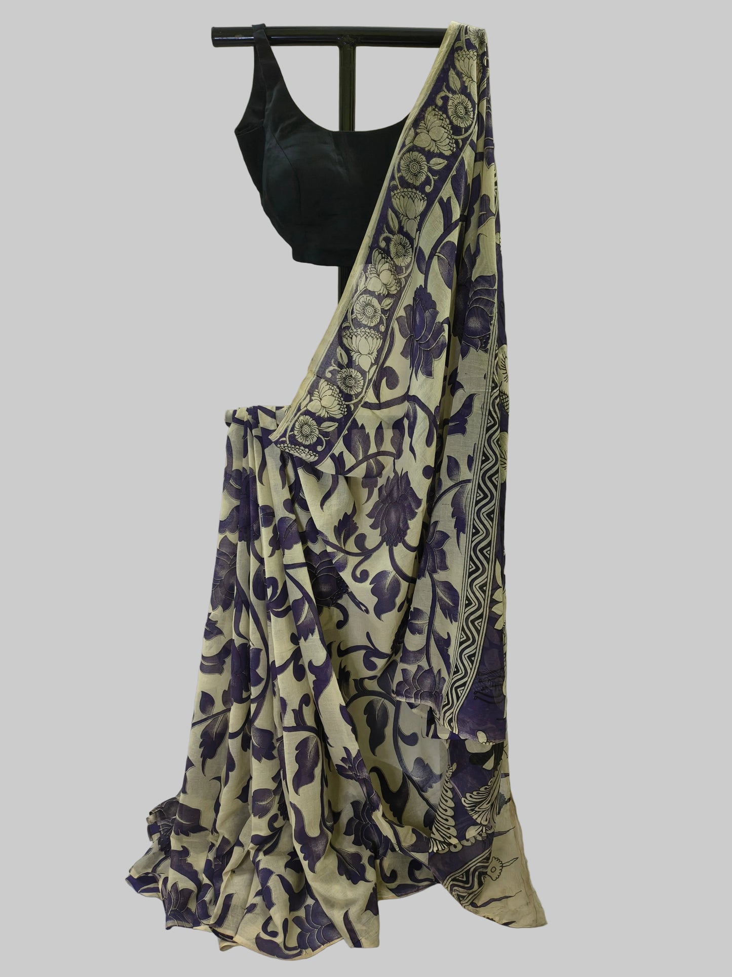 A beige and purple floral printed saree draped on a black hanger with a black sleeveless blouse, set against a draped off-white fabric backdrop.