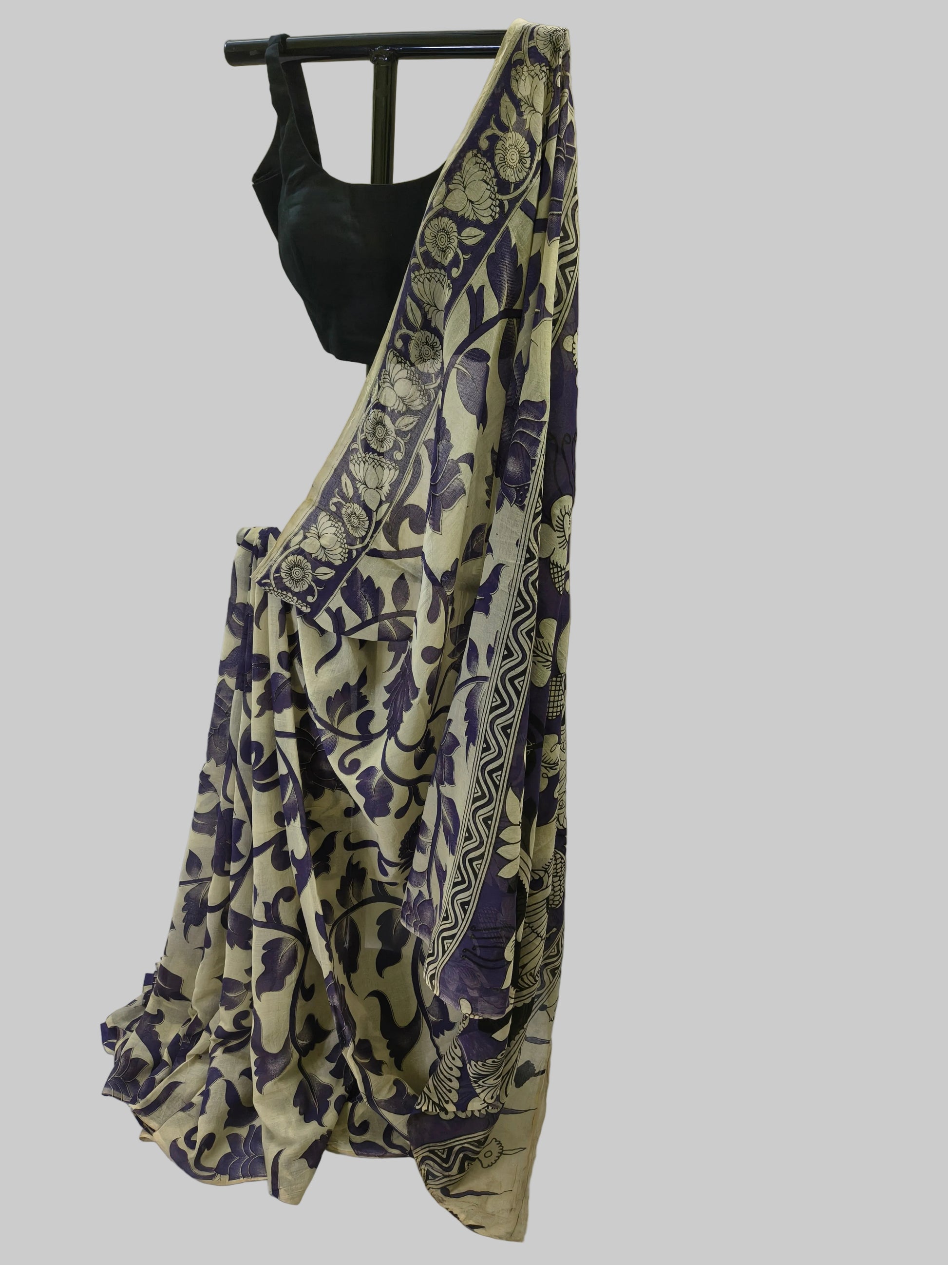 A beige and purple printed saree draped on a black hanger with a black blouse, set against a draped white fabric backdrop.