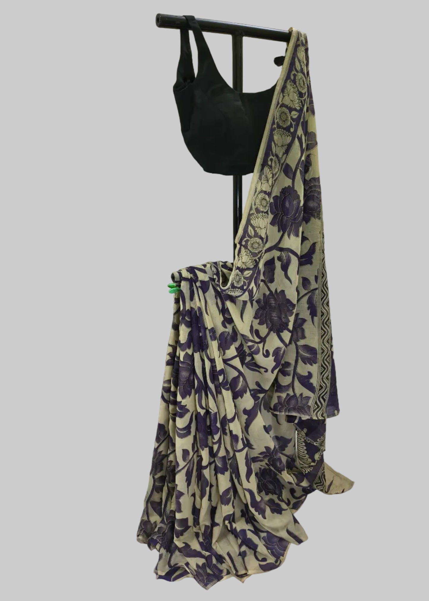 A beige saree with a dark purple floral and vine pattern, draped on a black dress form stand alongside a black blouse, set against a white fabric backdrop.