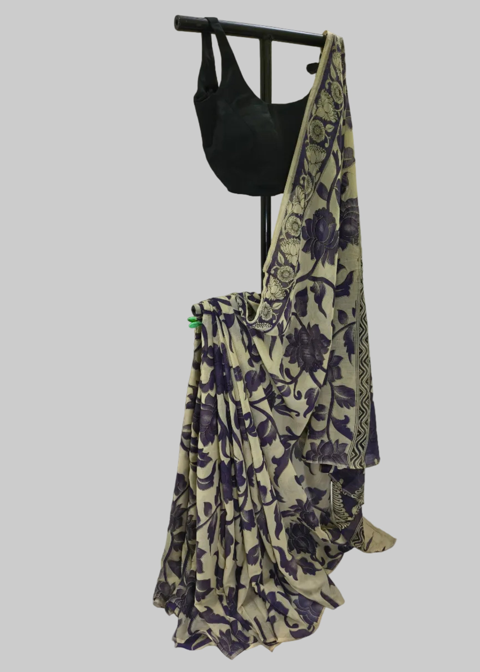 A beige saree with a dark purple floral and vine pattern, draped on a black dress form stand alongside a black blouse, set against a white fabric backdrop.