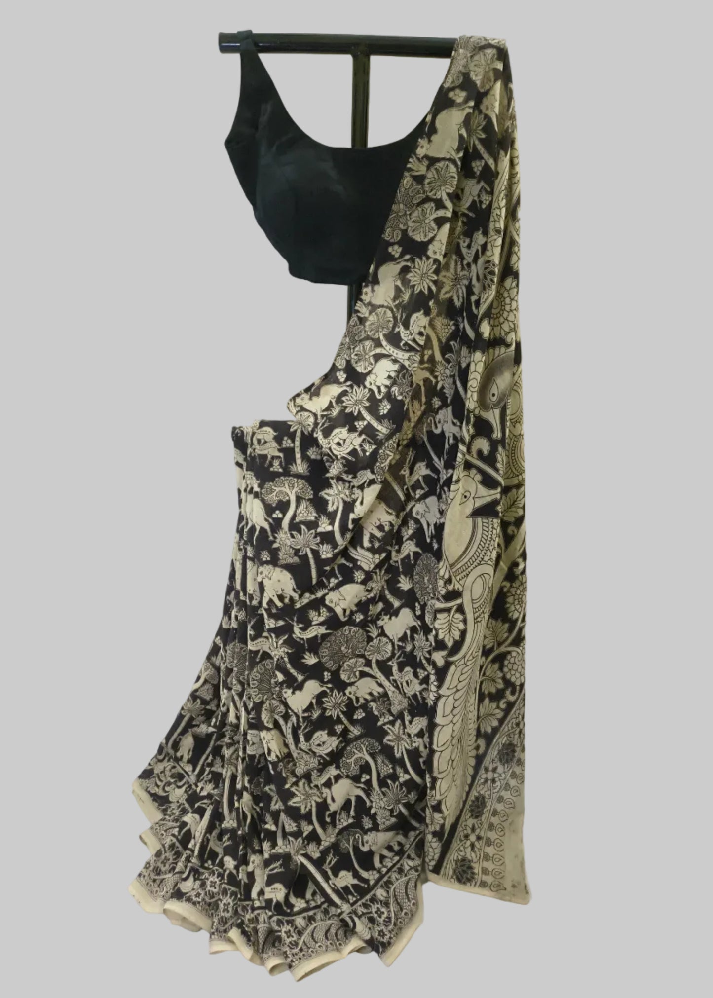A black and beige printed saree draped on a black dress form, paired with a solid black blouse, set against a draped off-white fabric backdrop.
