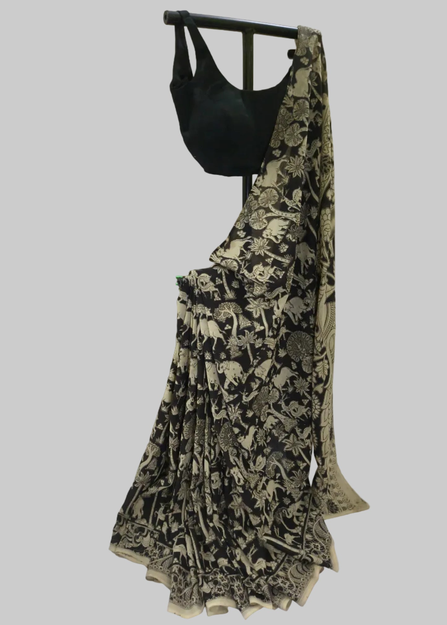 A black saree with beige animal and floral block prints, paired with a solid black blouse, displayed on a black clothing hanger against a draped white fabric backdrop.