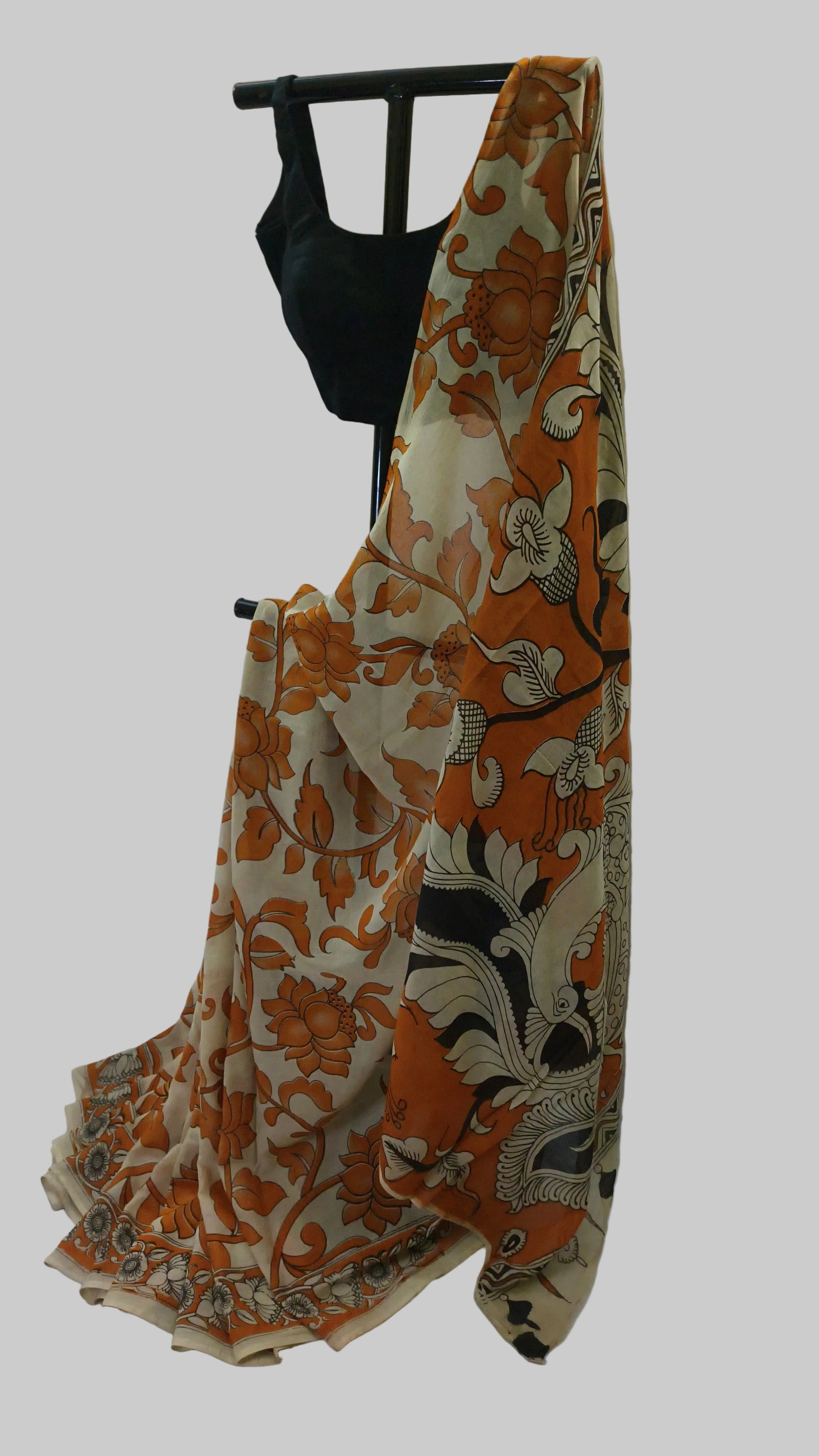 An orange, beige, and black floral printed saree draped over a black clothing hanger, paired with a black blouse, set against a white fabric backdrop.