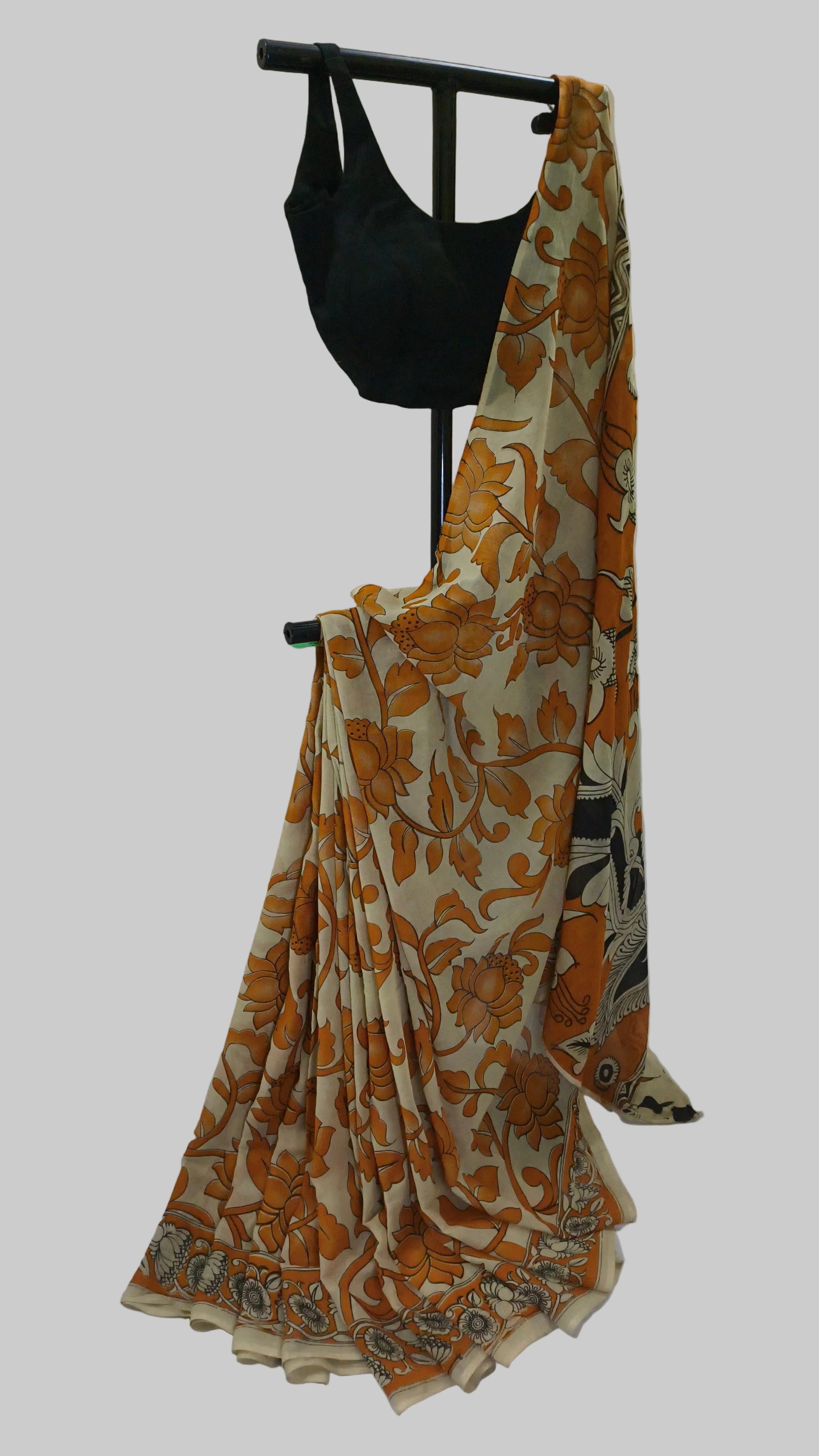 A beige and orange floral printed saree draped on a black clothing stand, paired with a solid black blouse, set against a plain white fabric backdrop.