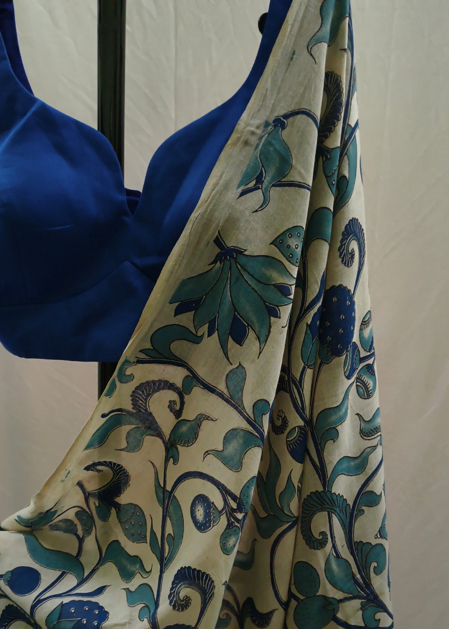 A cream-colored saree with a blue and teal floral block print, draped over a royal blue satin blouse on a black hanger against a white fabric backdrop.