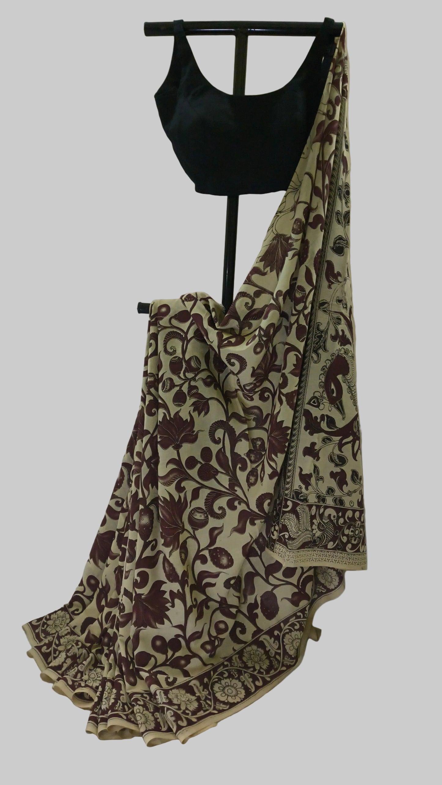 A beige and brown floral printed saree draped on a black dress form, paired with a black blouse, set against a draped white fabric backdrop.