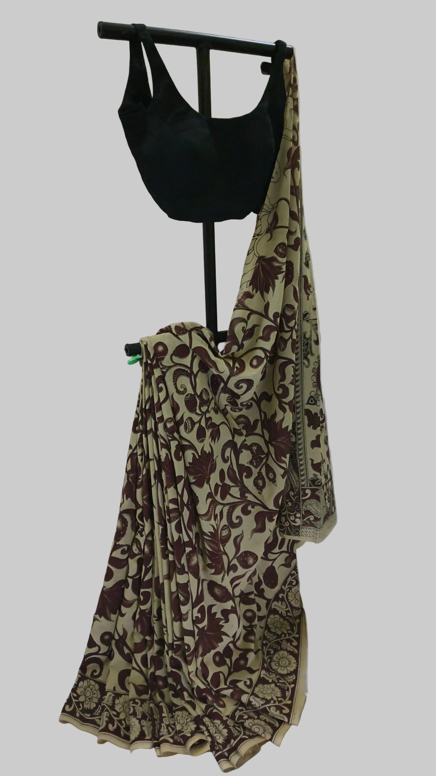 A beige saree with a dark brown floral vine pattern, draped on a black clothing stand alongside a black sleeveless blouse, set against a white fabric backdrop.
