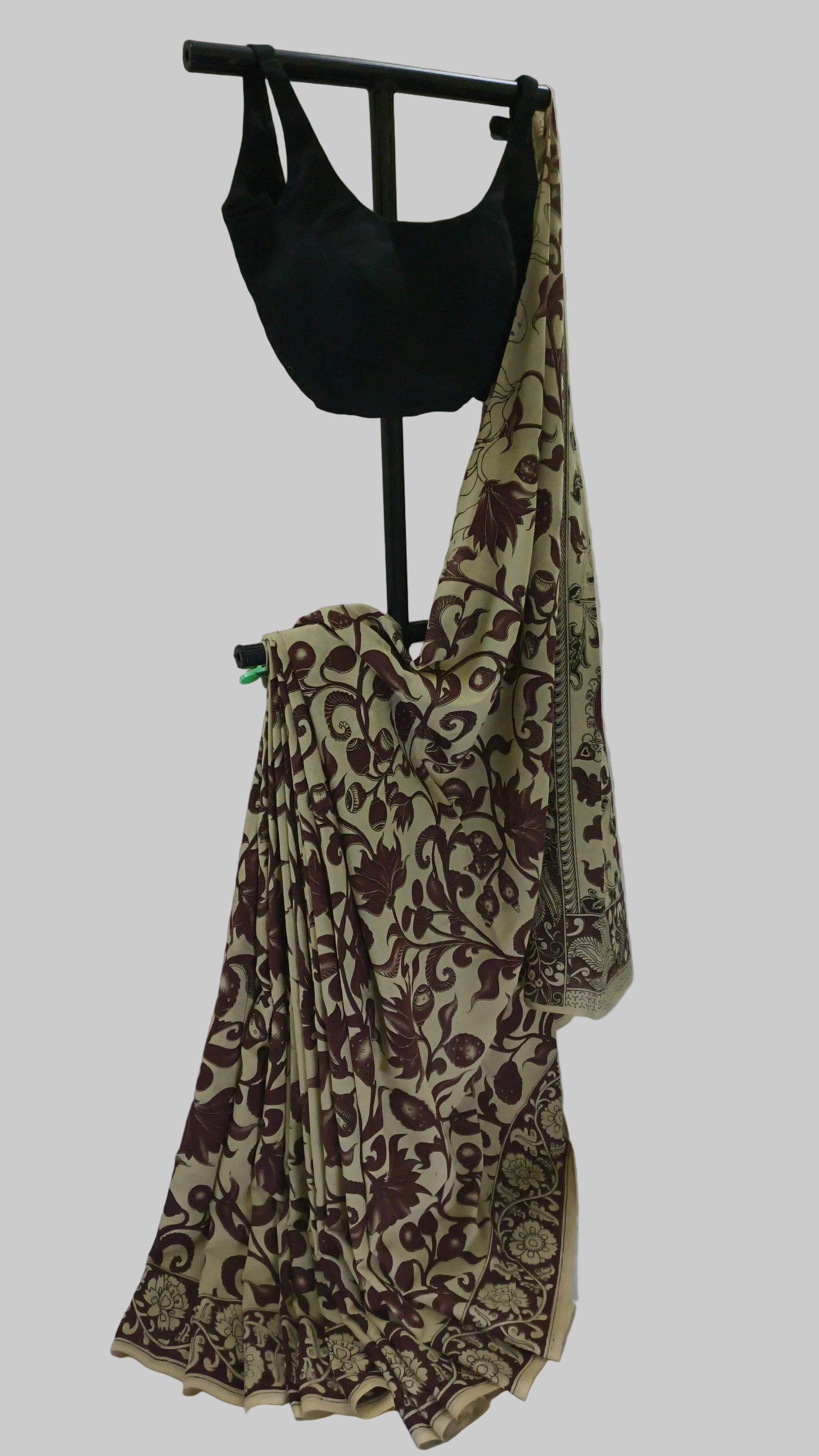 A beige saree with a dark brown floral vine pattern, draped on a black clothing stand alongside a black sleeveless blouse, set against a white fabric backdrop.
