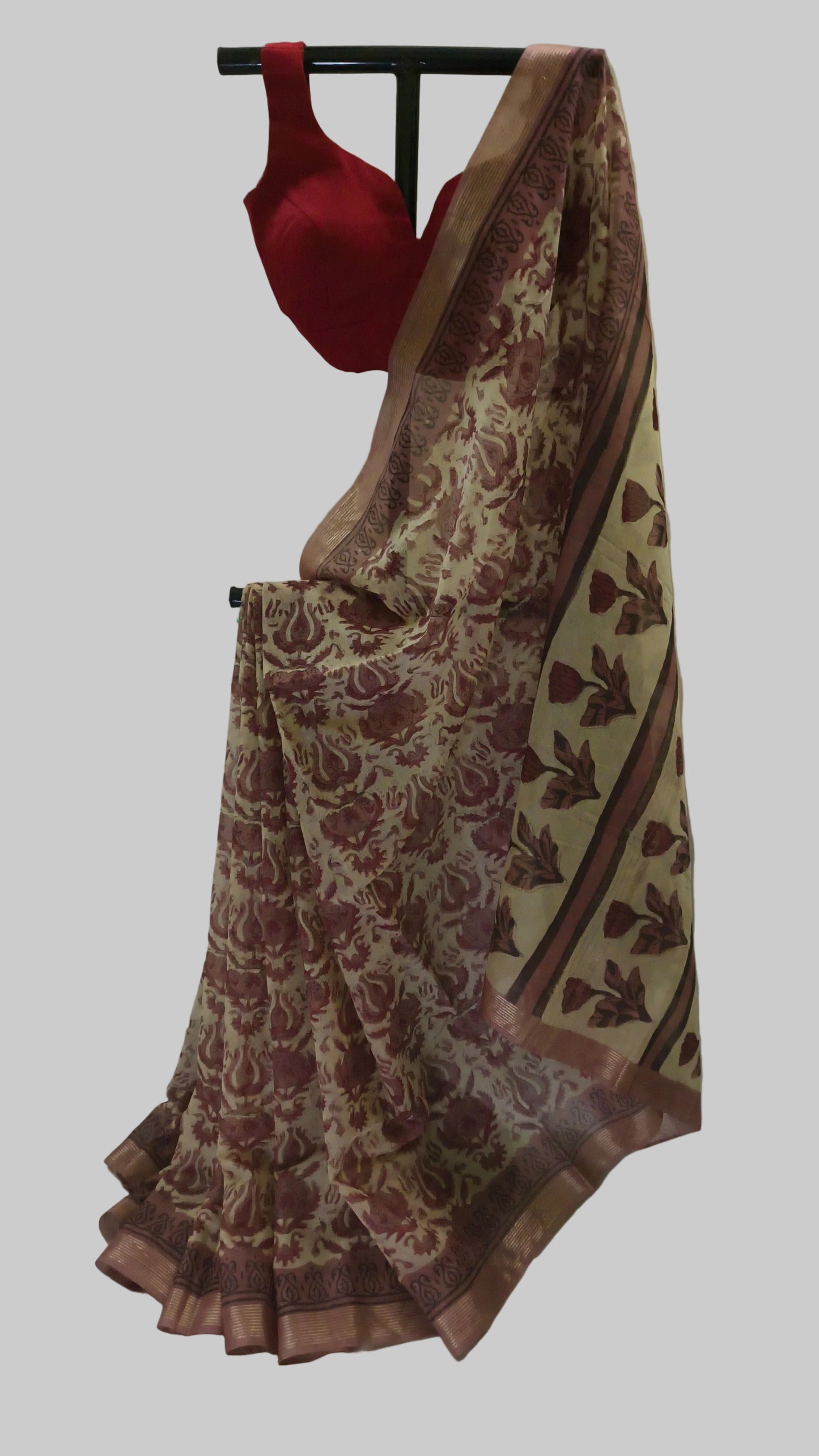 A beige and brown floral printed saree draped on a black hanger, paired with a solid red blouse, set against a draped white fabric backdrop.