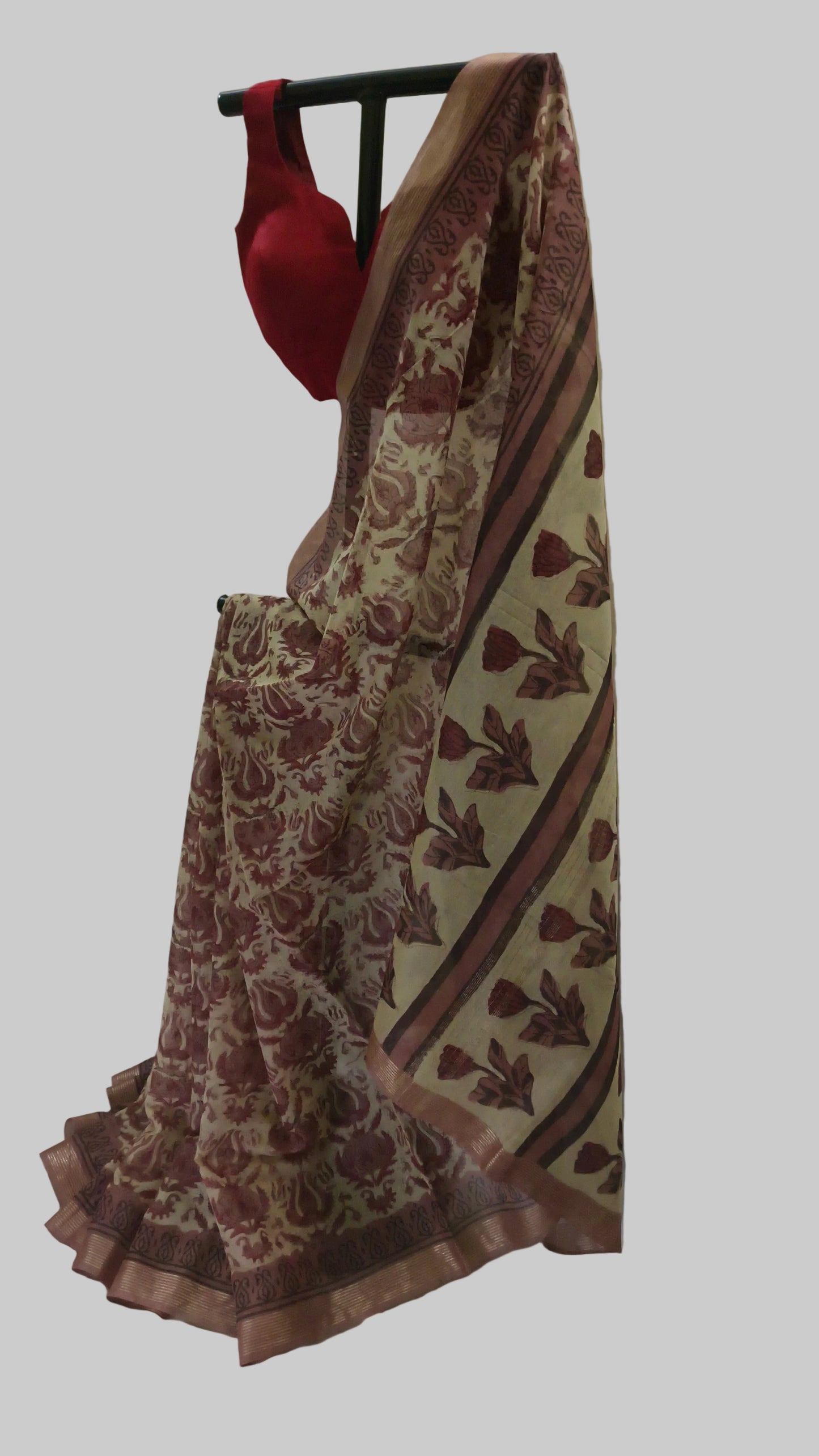 A beige and maroon printed saree draped on a black hanger with a red blouse, set against a draped white fabric backdrop.