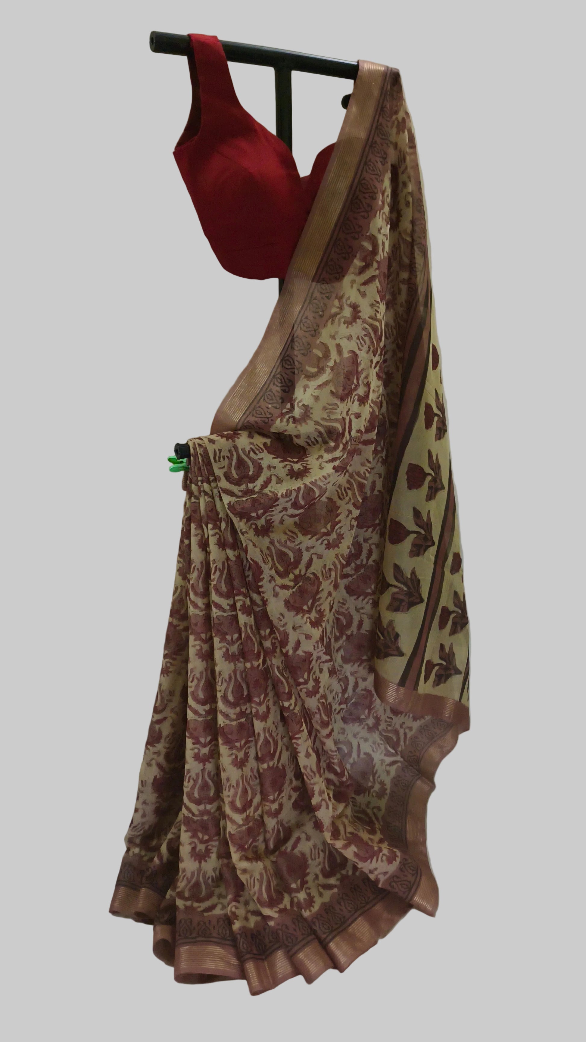 A beige and brown floral printed saree draped on a black hanger, paired with a solid red blouse, set against a draped white fabric backdrop.
