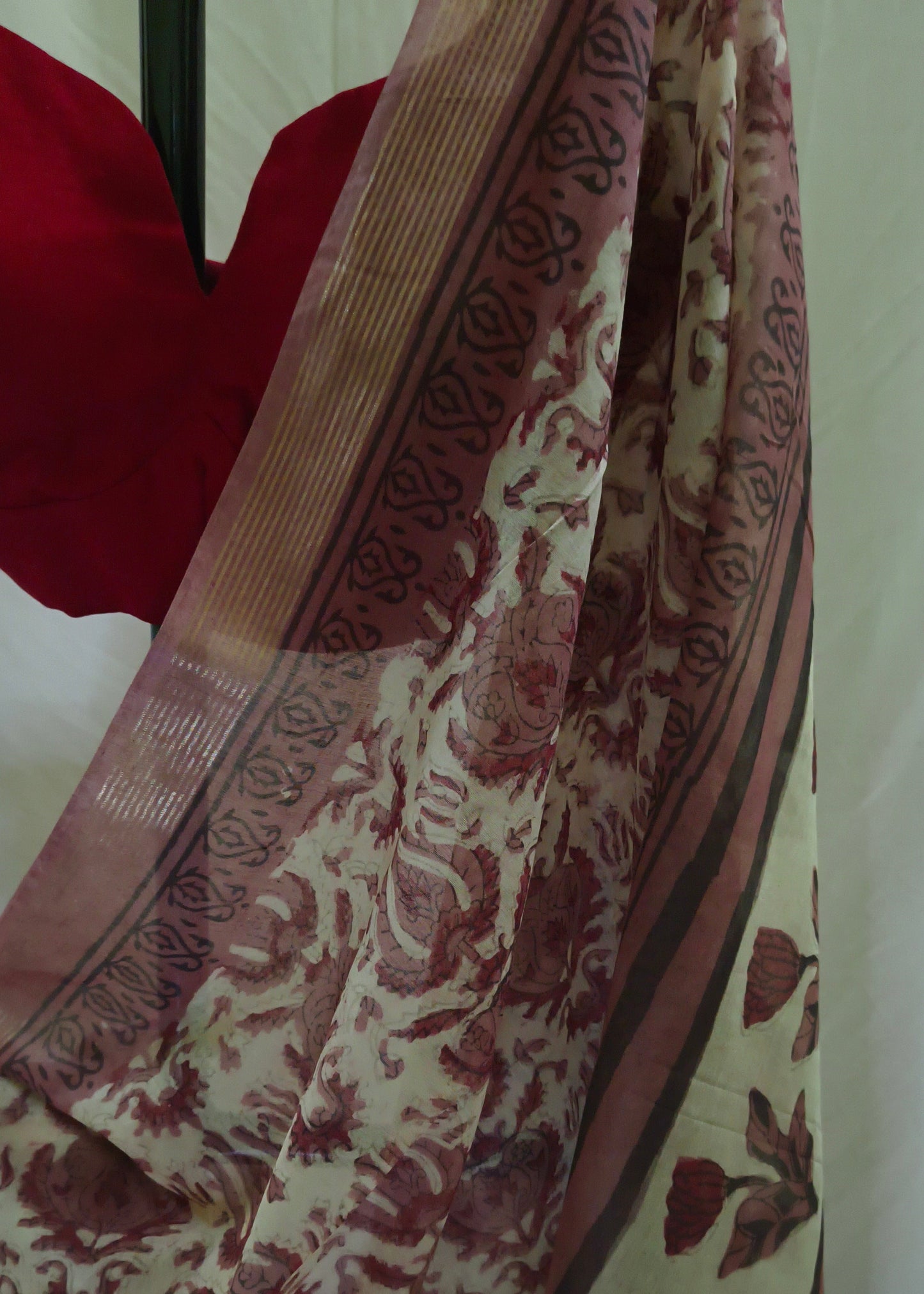 A close-up of a patterned saree or fabric draped over a mannequin, featuring a cream base with reddish-brown floral motifs and a matching red blouse visible at the top left.