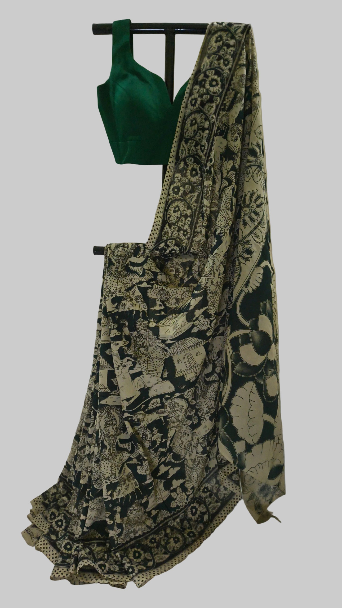 A black and beige printed saree draped over a black hanger, paired with a solid emerald green sleeveless blouse, set against a white fabric backdrop.