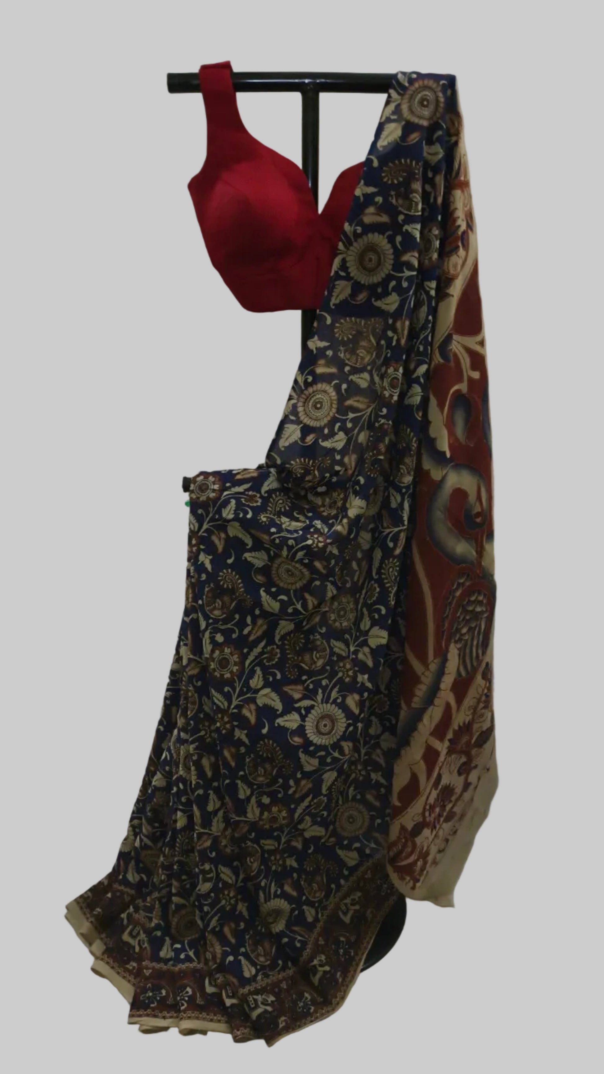 A traditional Indian saree displayed on a black hanger stand against a draped white fabric backdrop. The saree has a navy blue base with intricate beige and gold floral patterns, a red blouse, and a beige border with red and blue paisley motifs.
