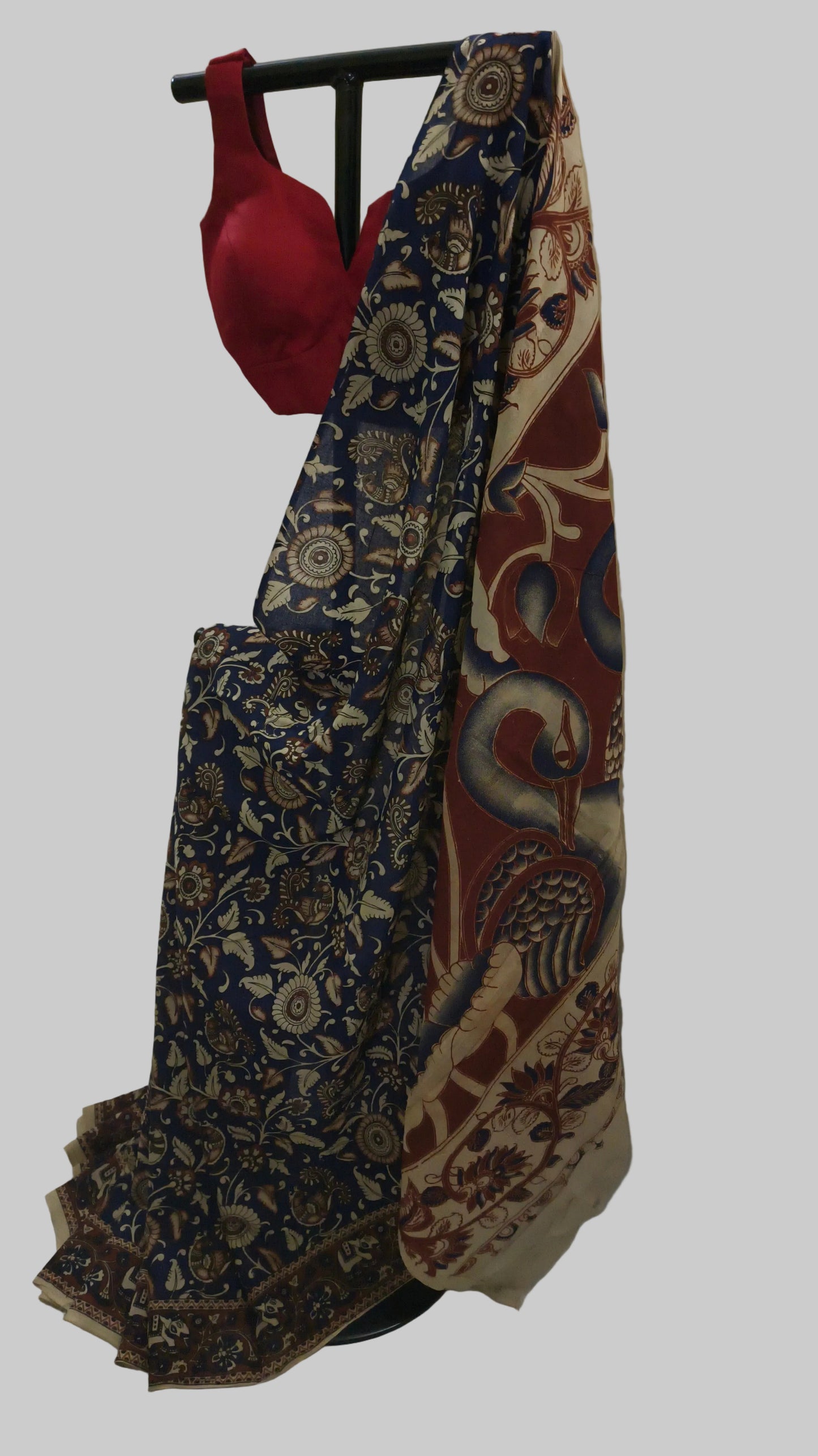 A traditional Indian saree draped on a black clothing stand, featuring a navy blue floral patterned body, a beige border with red and blue paisley motifs, and a solid red blouse attached at the top.