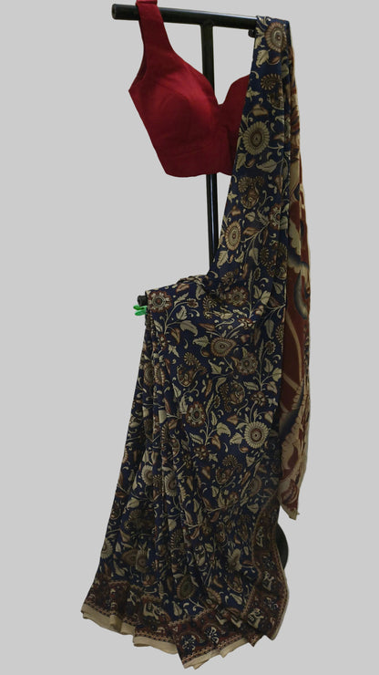 A traditional Indian saree displayed on a black stand against a white fabric backdrop. The saree features a navy blue base with intricate beige and brown floral patterns, paired with a solid red blouse.