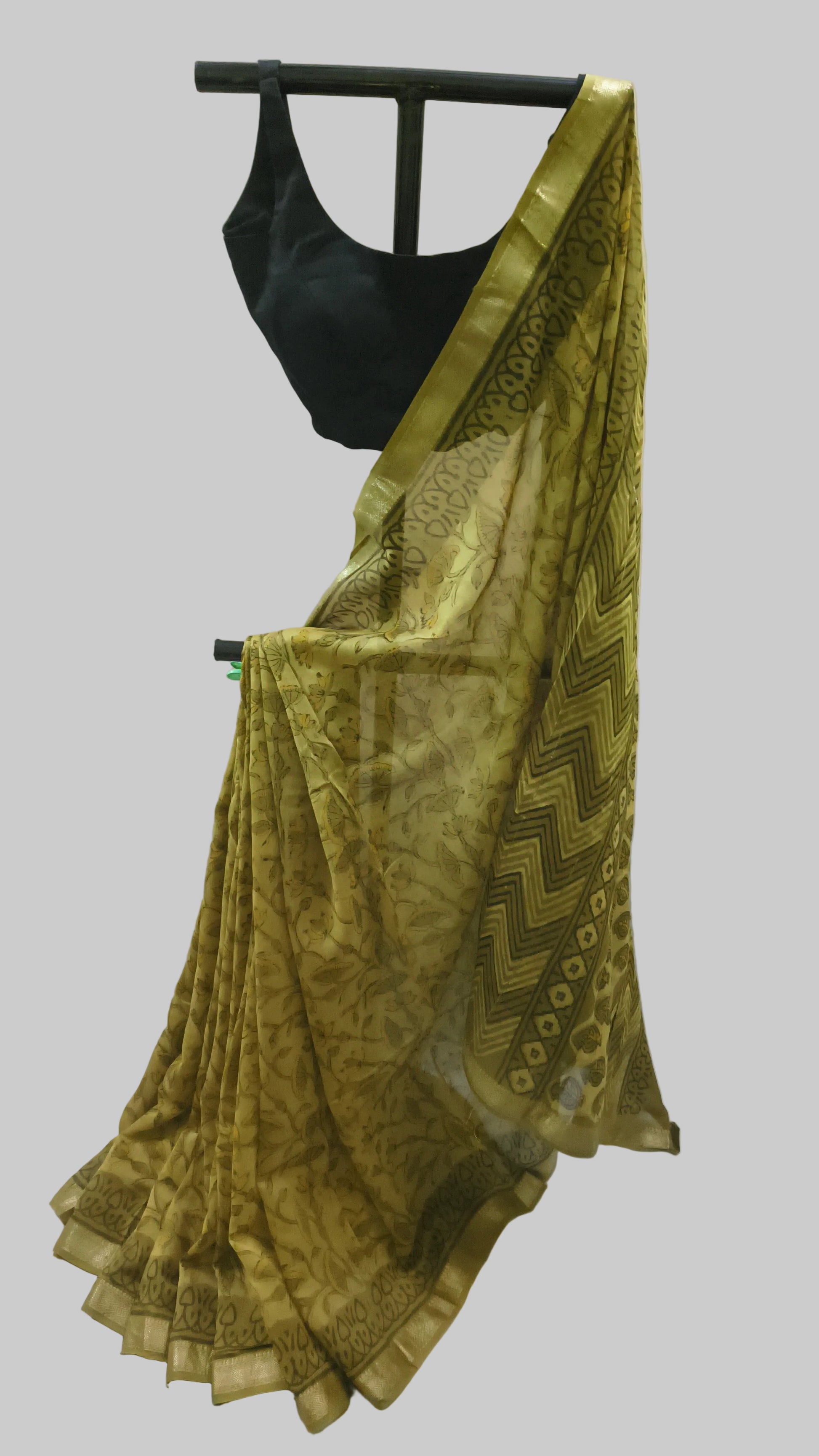 A yellow-gold patterned saree with a black blouse displayed on a black hanger against a white fabric backdrop.