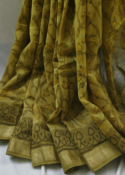 A draped olive green saree with a black floral print and a wide gold zari border, shown against a white fabric backdrop.