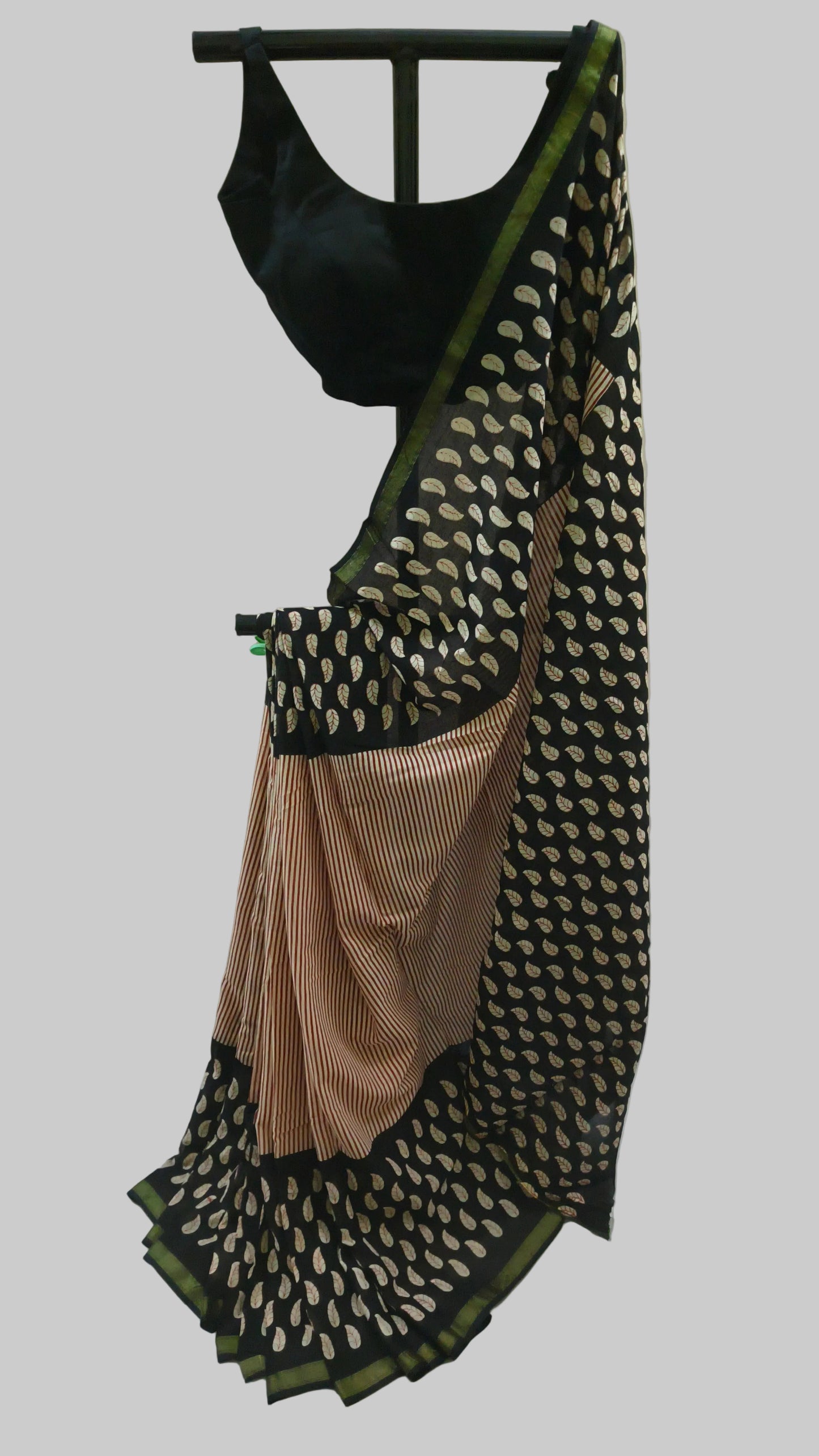 A black and beige saree with a green border, draped on a black hanger against a white fabric backdrop. The saree features a black blouse with a scoop neck, and the body is patterned with beige motifs on a black base, with a solid beige pleated section at the pallu.