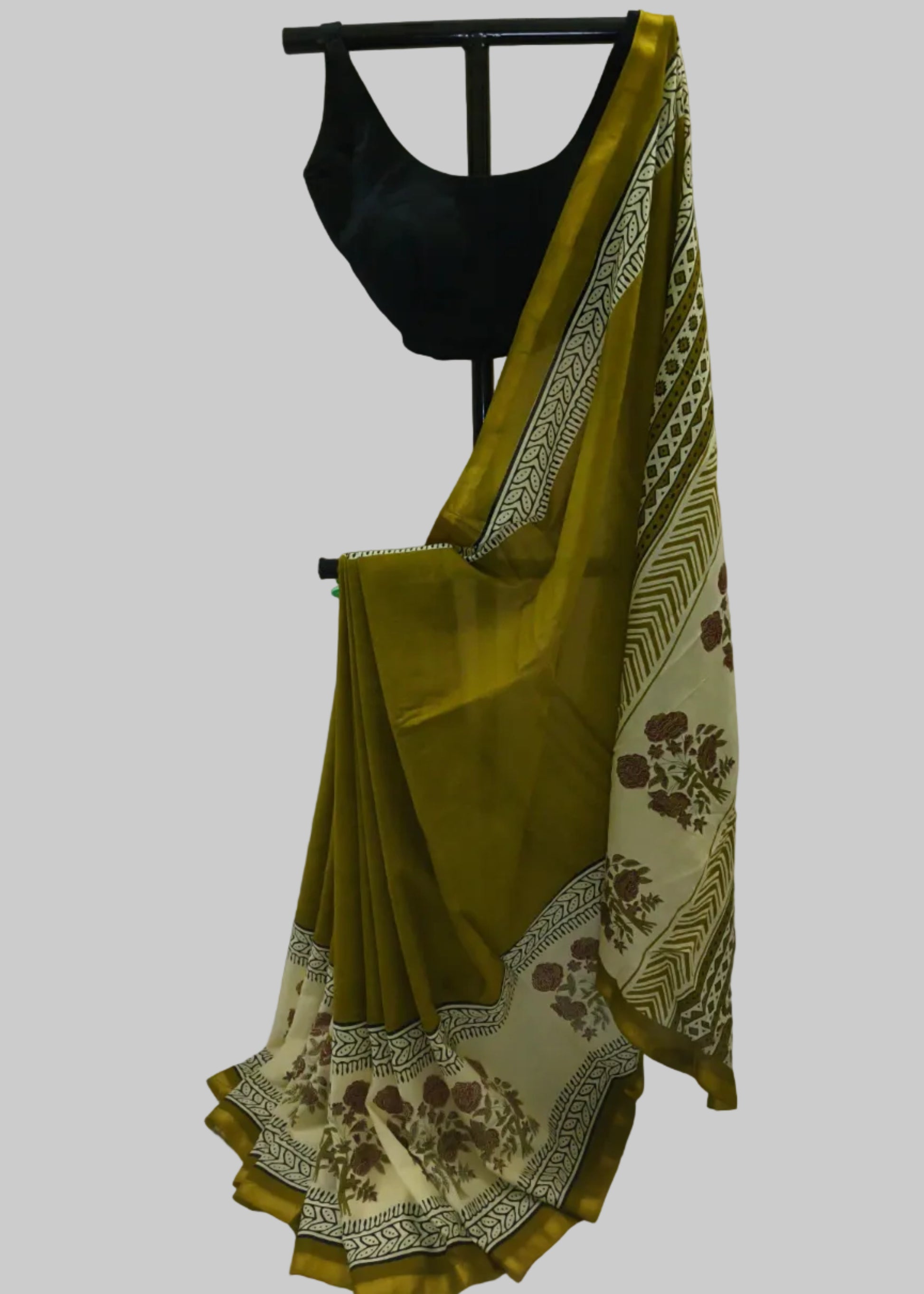 An olive green saree with a cream and brown floral border, draped on a black hanger stand with a black blouse, set against a white fabric backdrop.