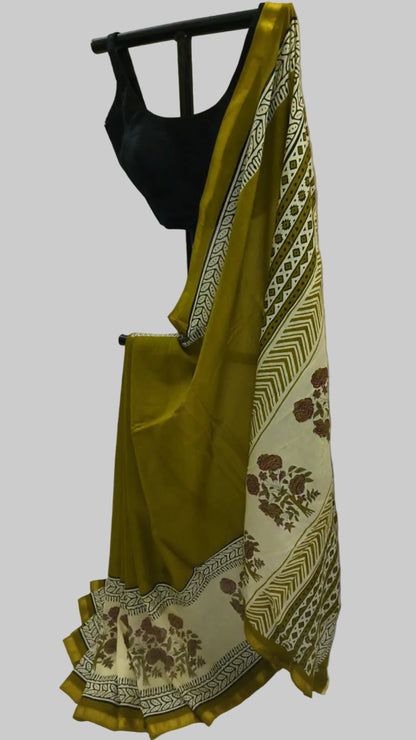 An olive green saree with a cream and brown patterned border, draped on a black hanger with a black blouse, set against a white fabric backdrop.