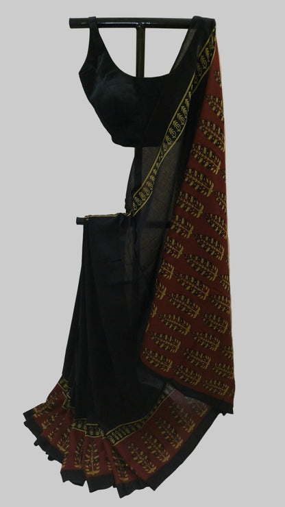 A black cotton saree with a patterned maroon border and a matching black blouse, displayed on a hanger against a white fabric backdrop.