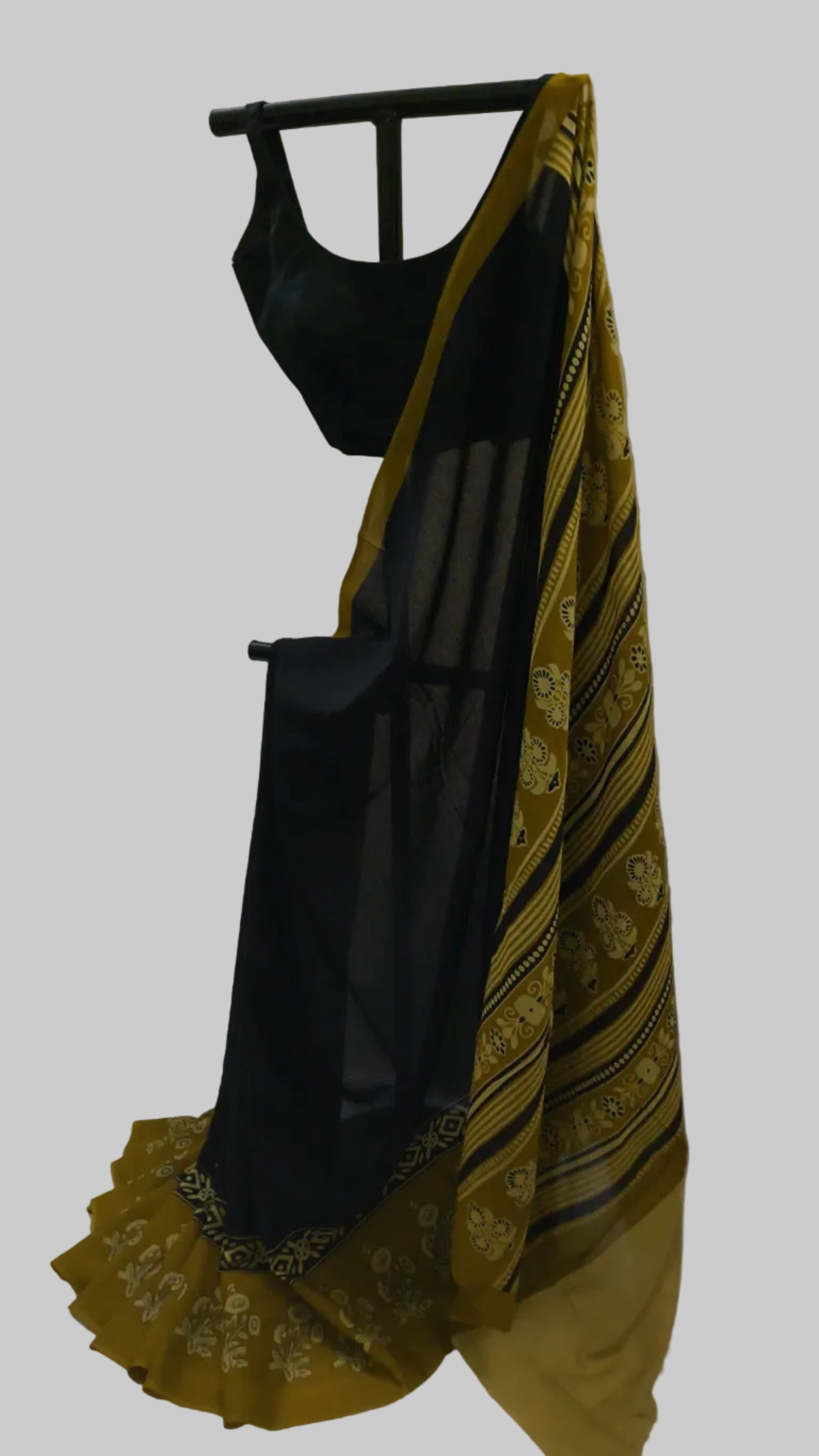 A black sleeveless blouse with a black saree featuring a mustard yellow border and gold floral patterns, displayed on a black hanger against a white fabric backdrop.