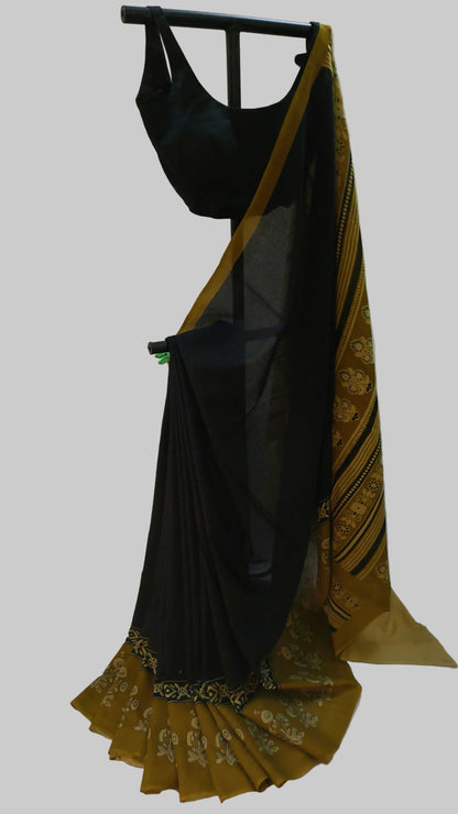 A black saree with a mustard yellow and black patterned border and pallu, displayed on a black hanger against a draped white fabric backdrop.