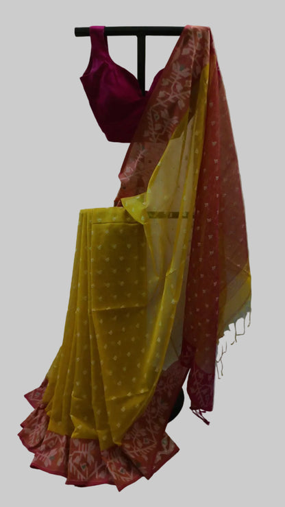 A traditional Indian saree displayed on a black clothing rack against a white draped backdrop. The saree features a bright yellow body with small white motifs, a deep red border, and a magenta blouse.