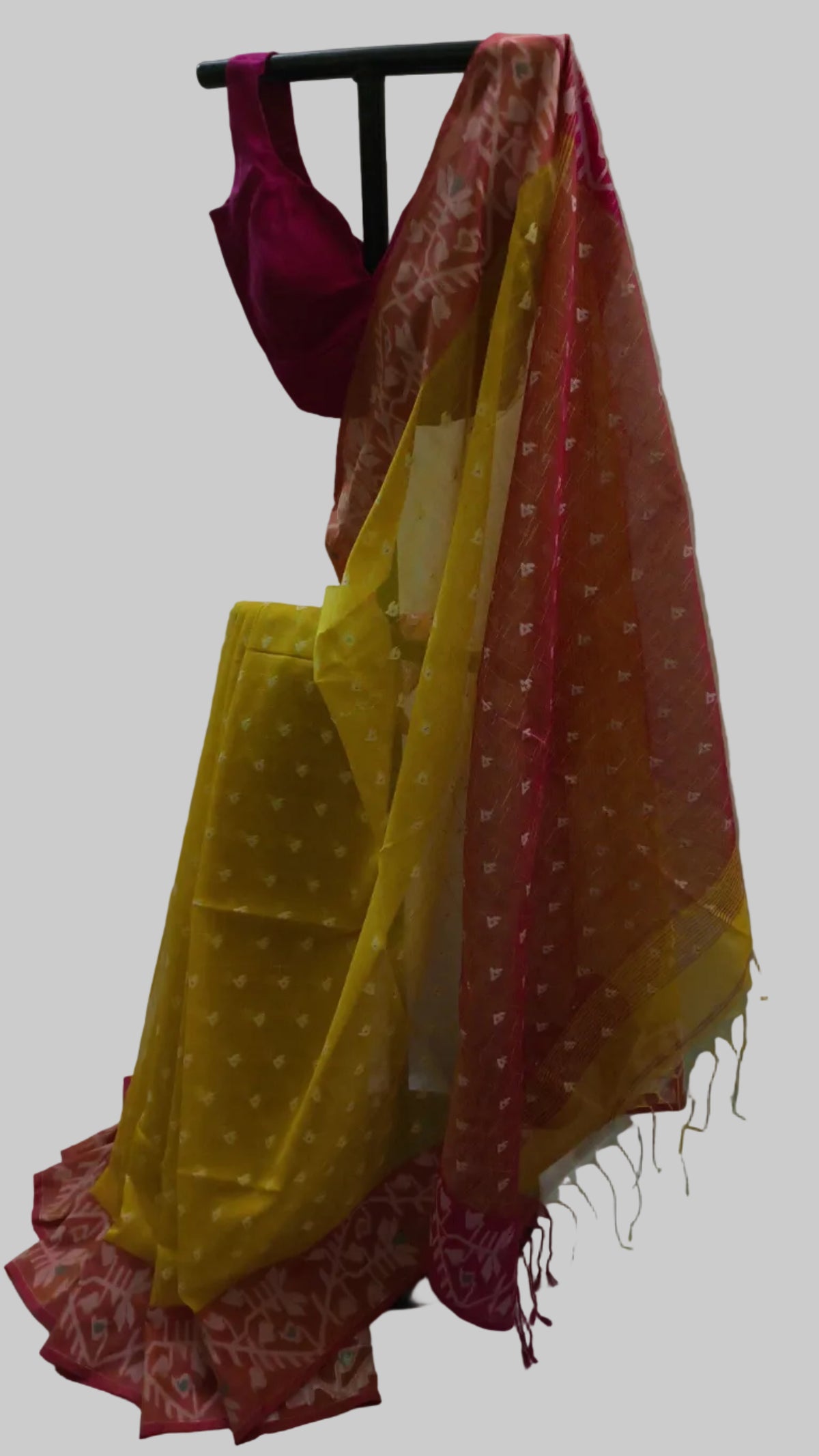 A traditional Indian saree displayed on a black clothing rack against a white fabric backdrop. The saree features a bright yellow body with white polka dots, a pinkish-red border with white motifs, and a matching magenta blouse.