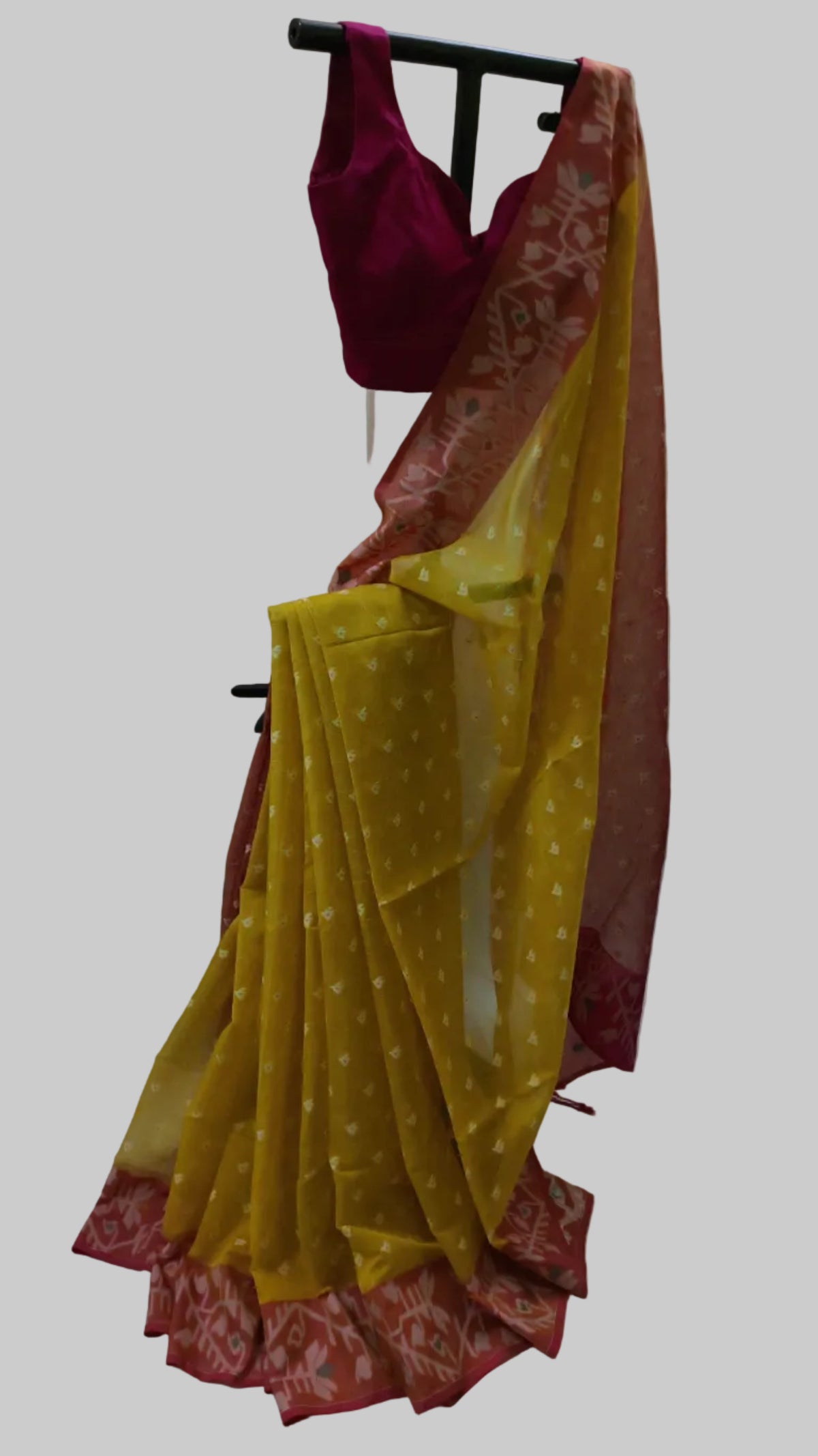A yellow and red saree draped on a black clothing rack, featuring a deep magenta blouse, with a white draped fabric background.