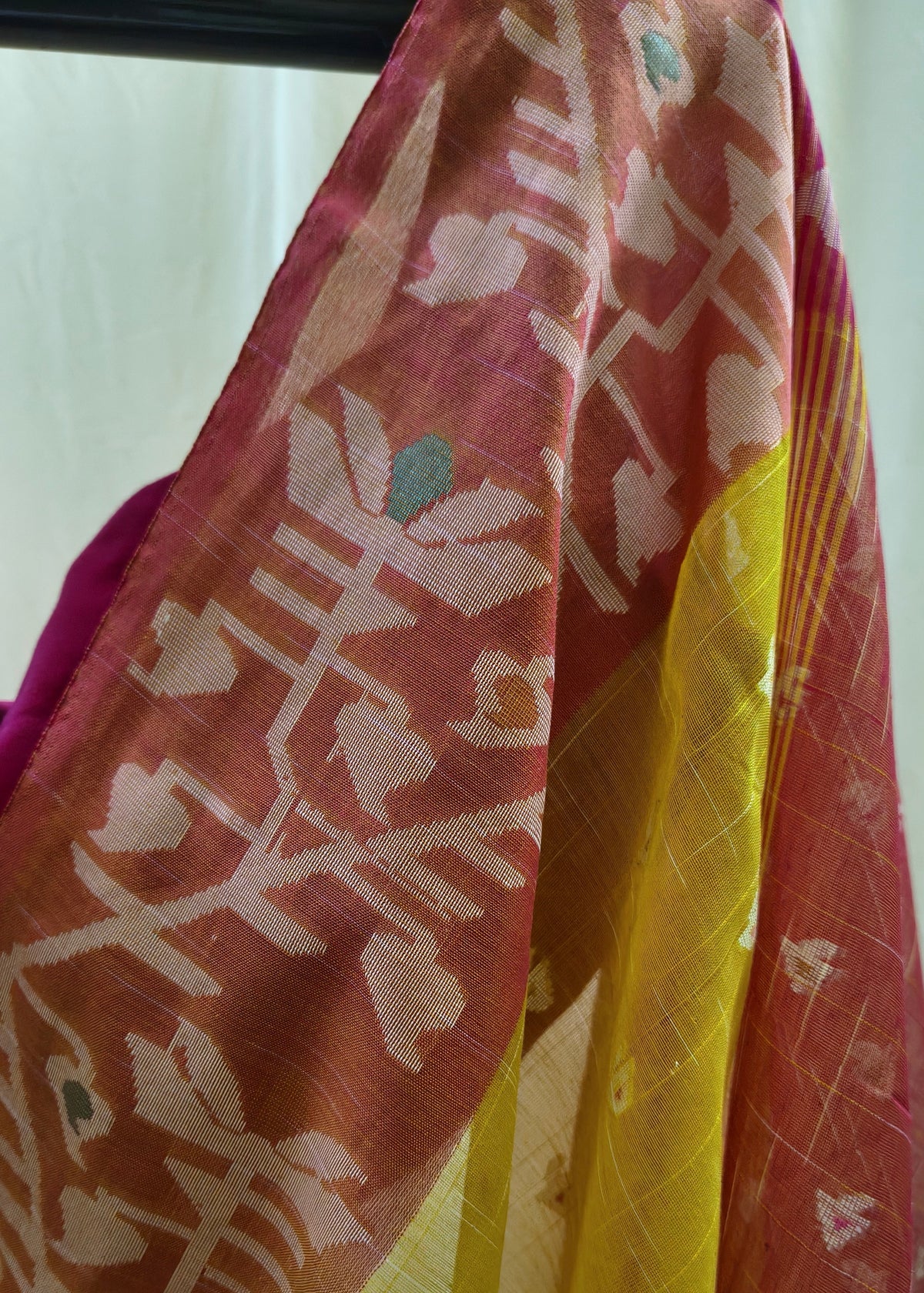 A close-up of a draped patterned fabric, likely a saree or scarf, featuring a rust-red base with white floral motifs and a prominent yellow border.