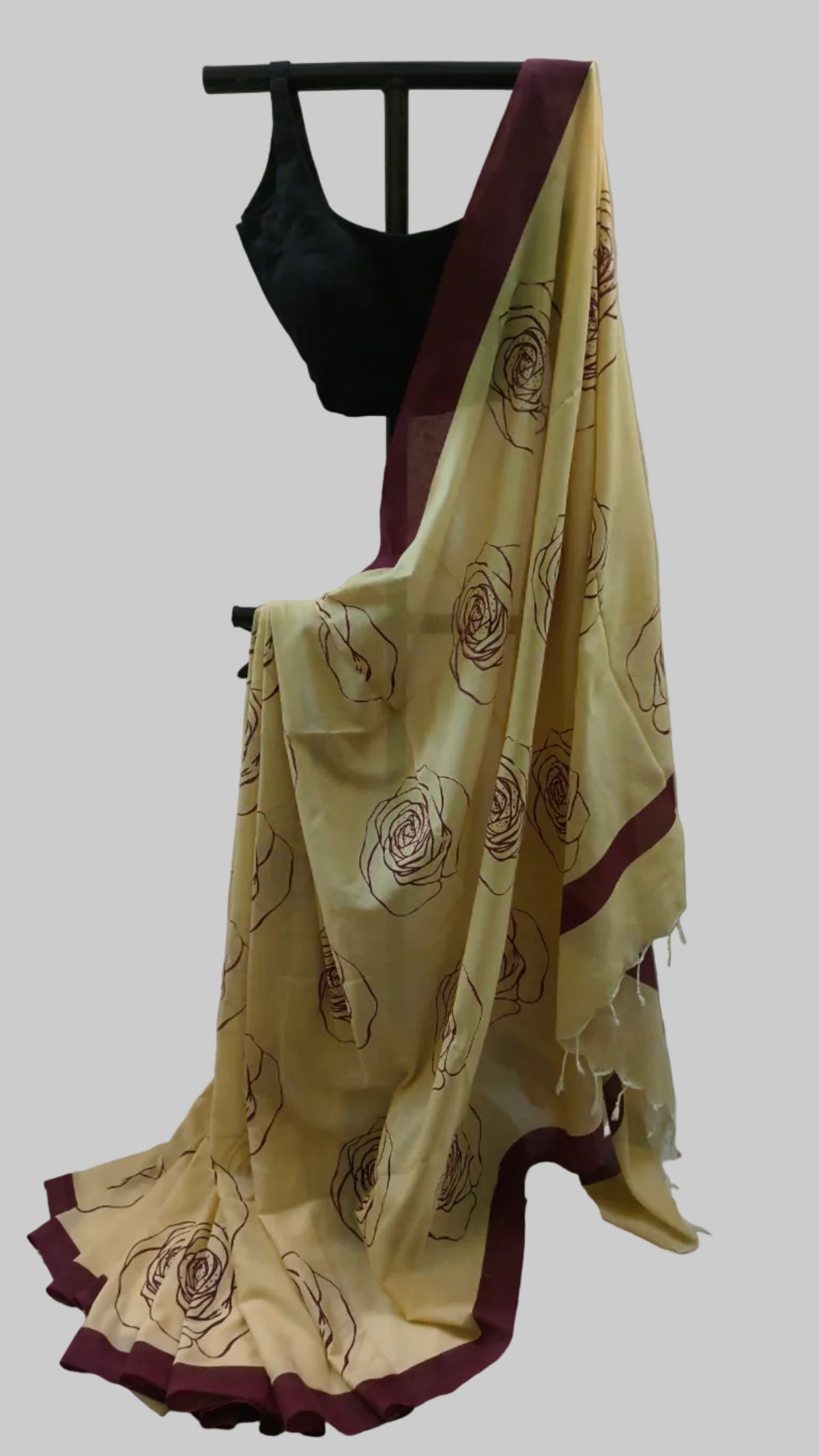 A cream-colored saree with dark brown floral patterns and deep maroon borders, paired with a black blouse, displayed on a black hanger against a neutral draped backdrop.