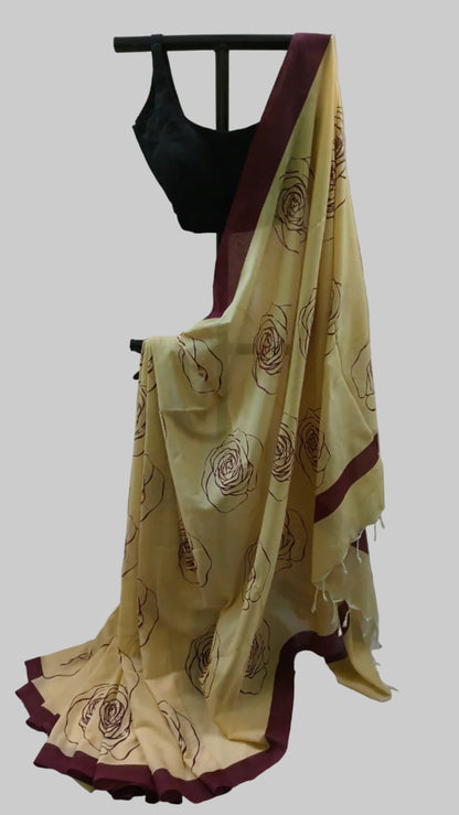 A cream-colored saree with dark brown floral patterns and deep maroon borders, paired with a black blouse, displayed on a black hanger against a neutral draped backdrop.